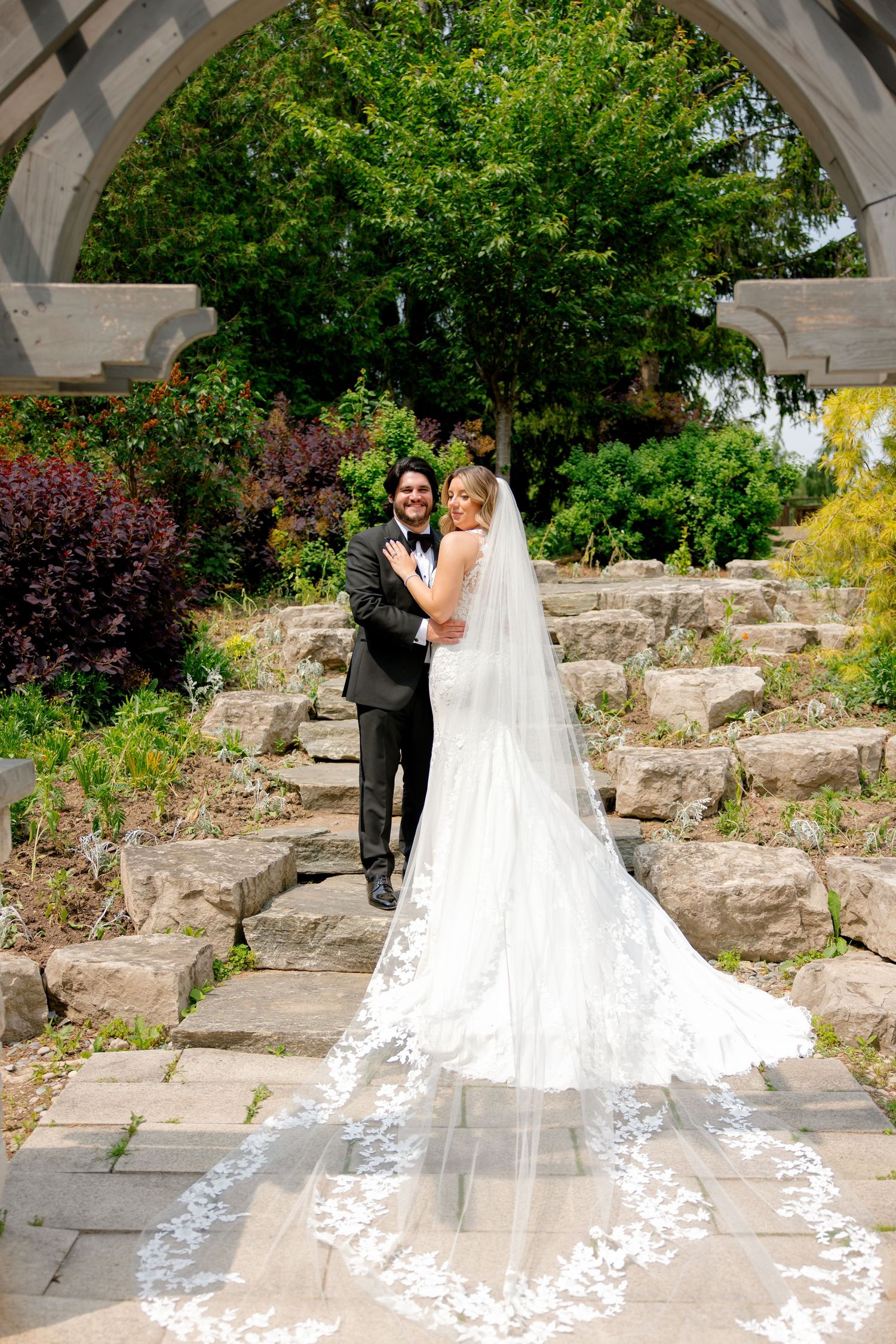 Wedding couple poses under stone archway with dramatic cathedral veil trailing down stone steps.