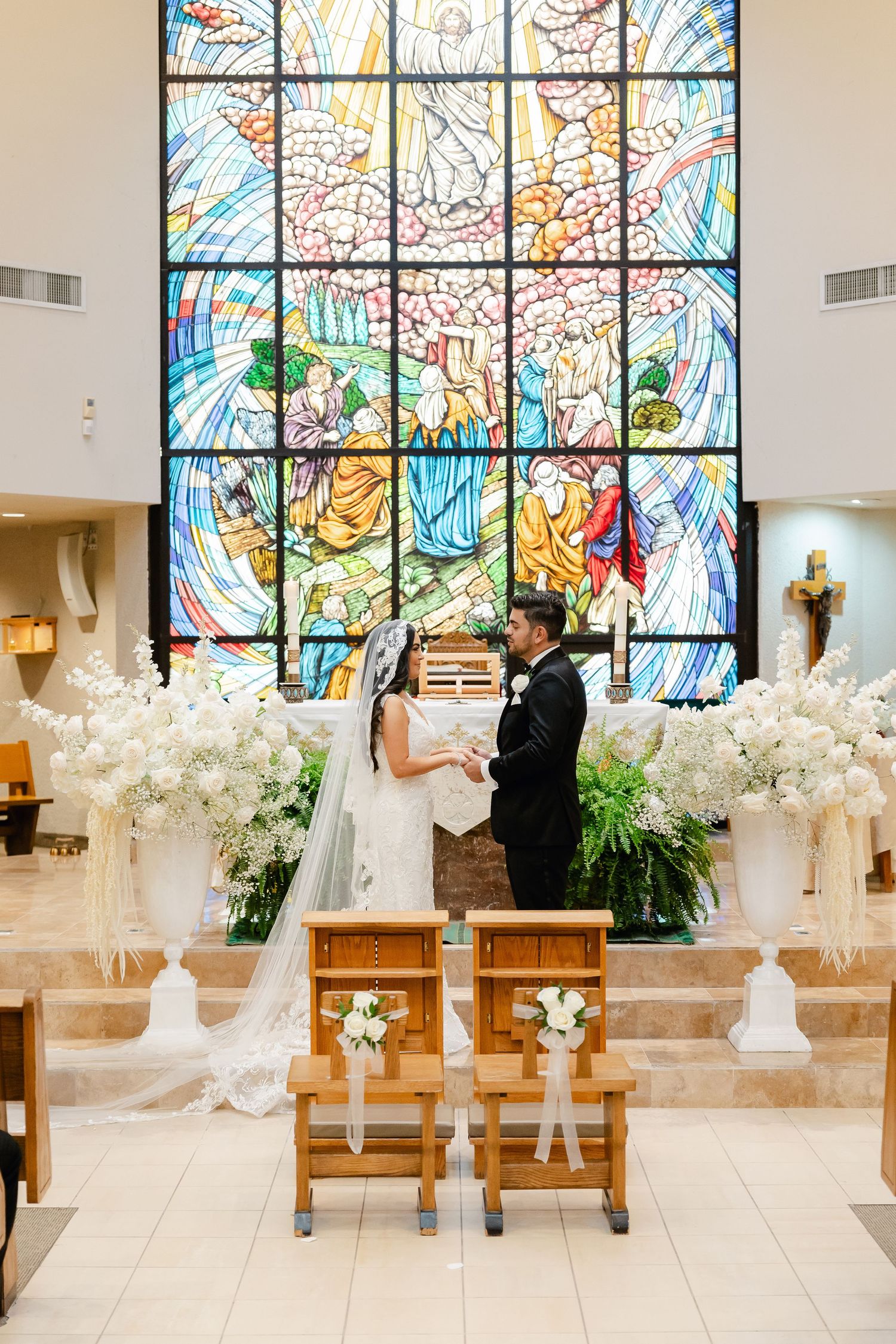 Wedding ceremony with couple before colorful stained glass window and white floral arrangements.
