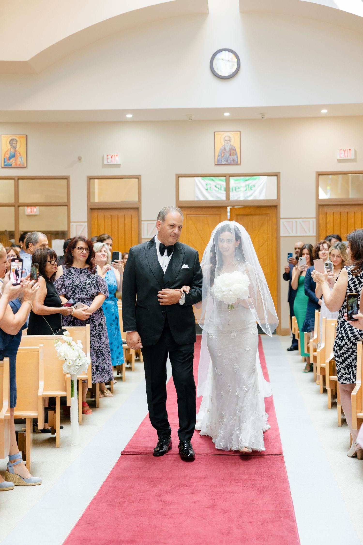 Bride in a lace gown walks down the red-carpeted church aisle with her escort as guests look on.