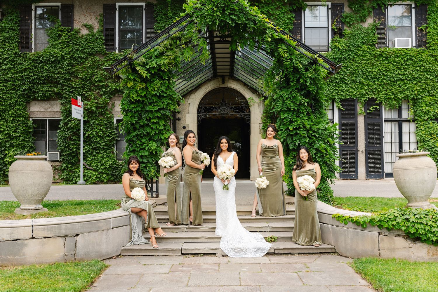 Wedding party with bridesmaids in taupe dresses poses at entrance of ivy-covered venue.
