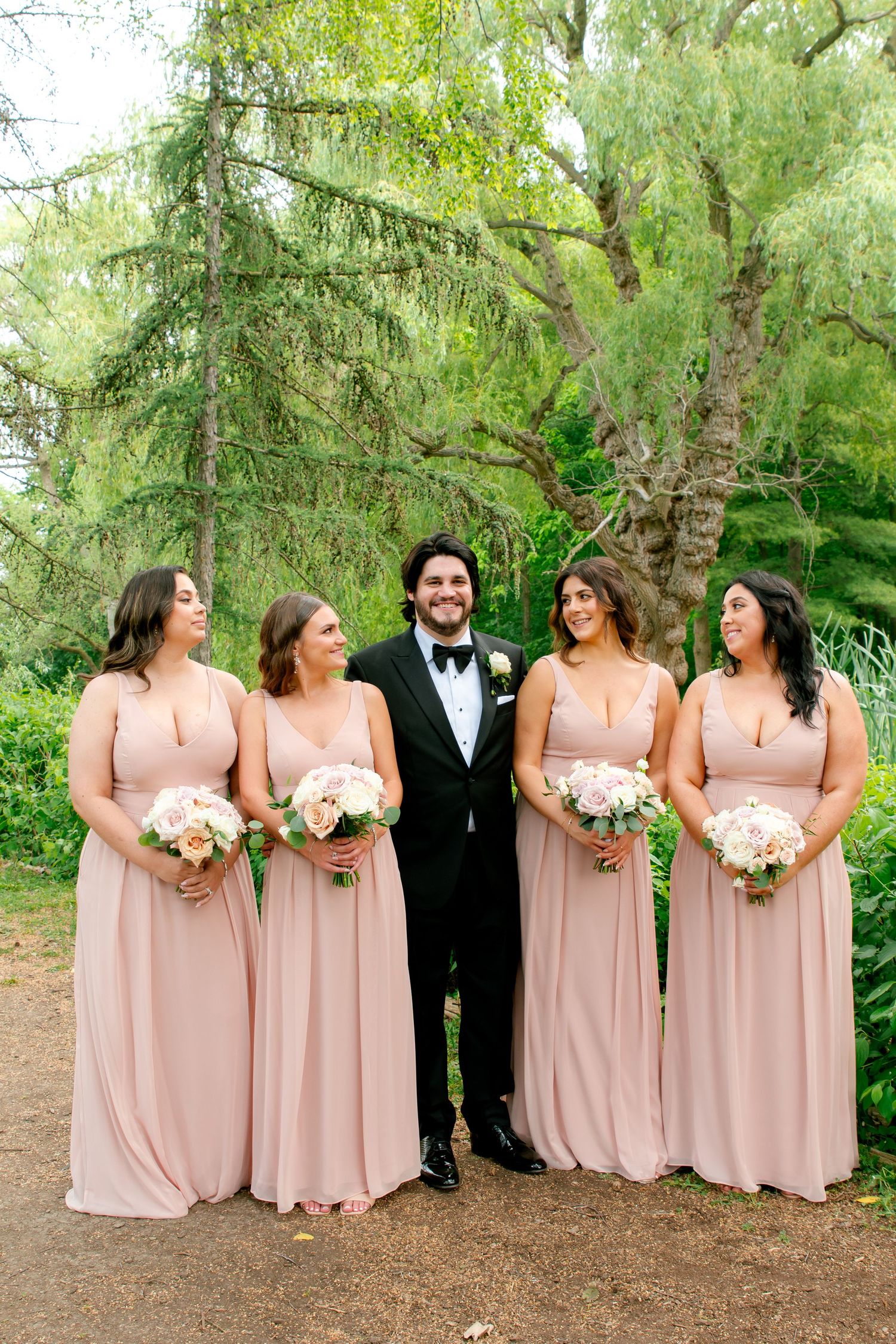 Bridal party standing together outdoors with bridesmaids in blush gowns holding white bouquets.