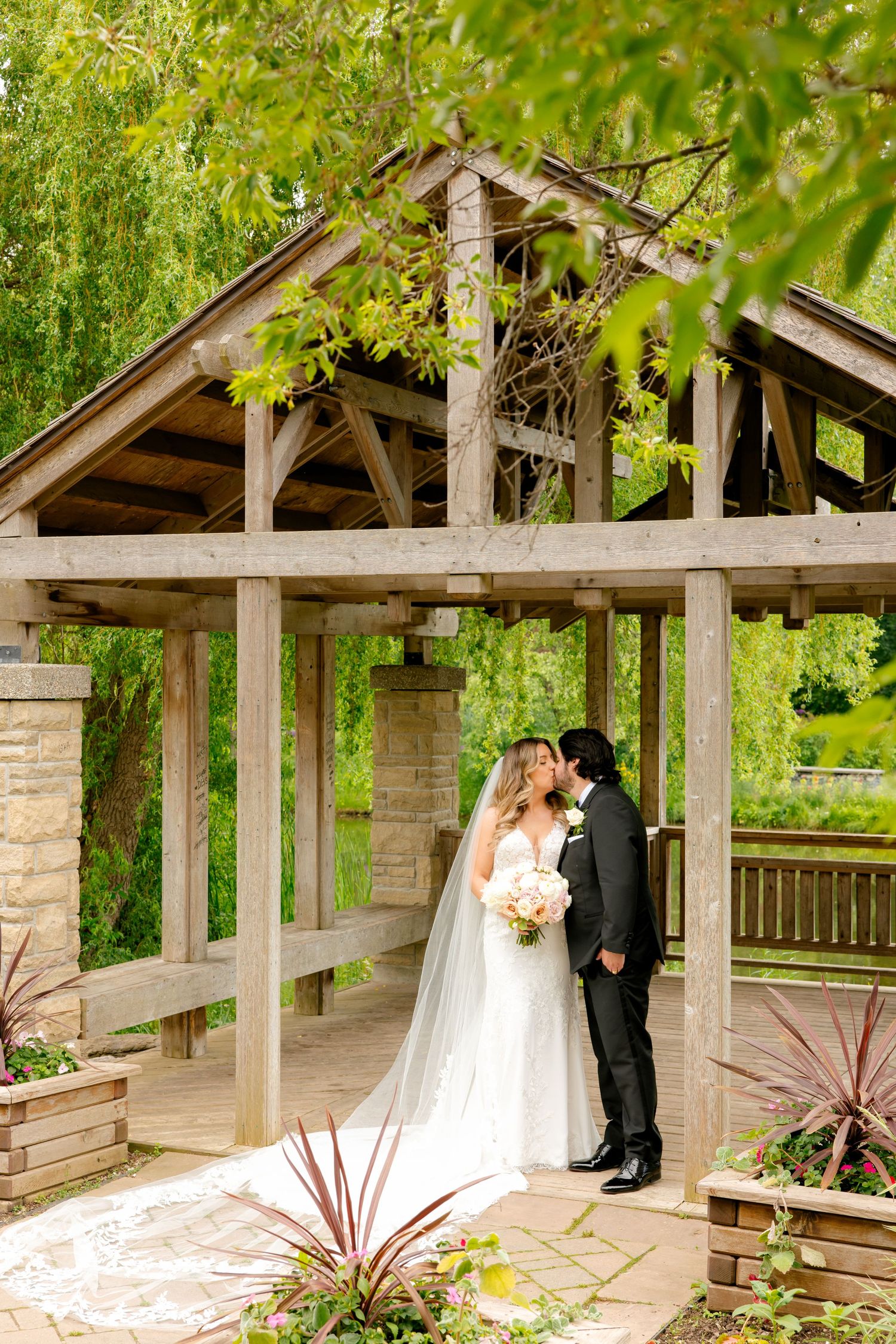 A couple shares an intimate moment under a rustic wooden gazebo surrounded by lush greenery.