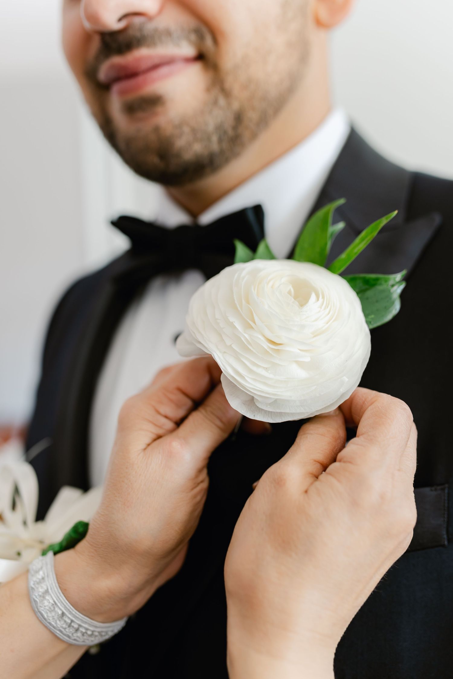 Close-up of hands pinning an elegant white ranunculus boutonniere on a black tuxedo.
