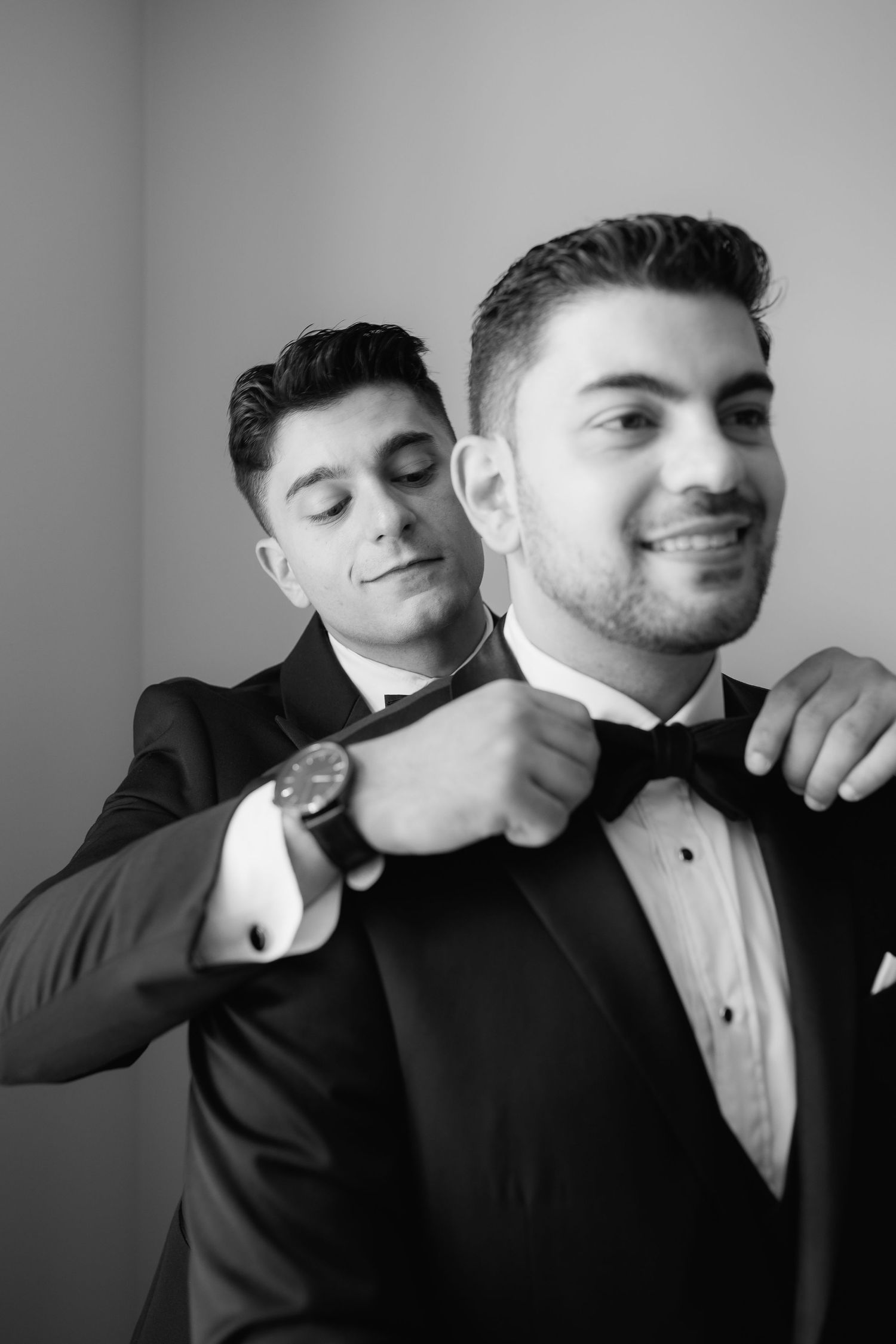Two people in tuxedos share a candid black and white moment while adjusting formal wear.