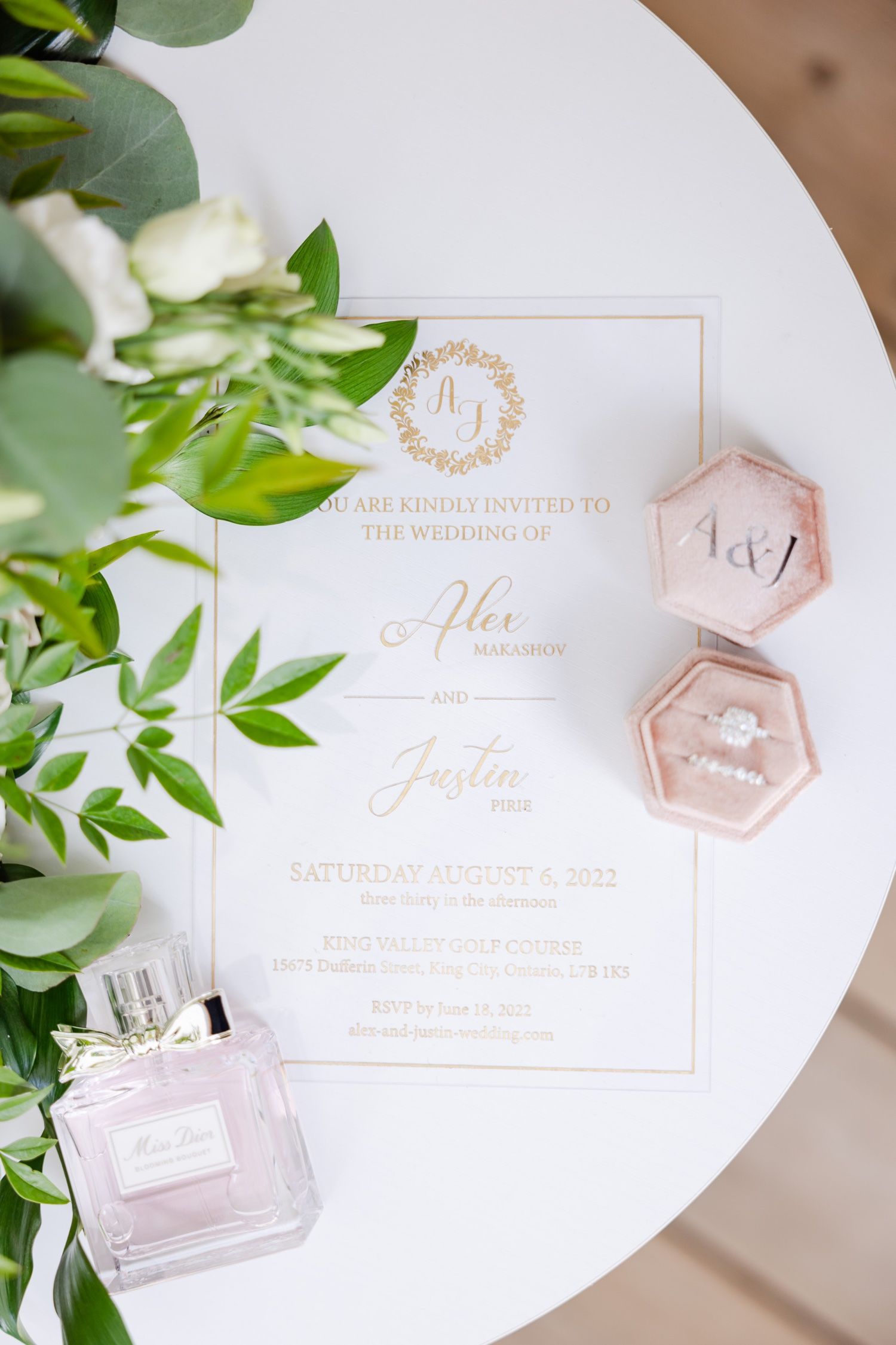 Wedding invitation with gold text alongside pink ring box and perfume on white table.