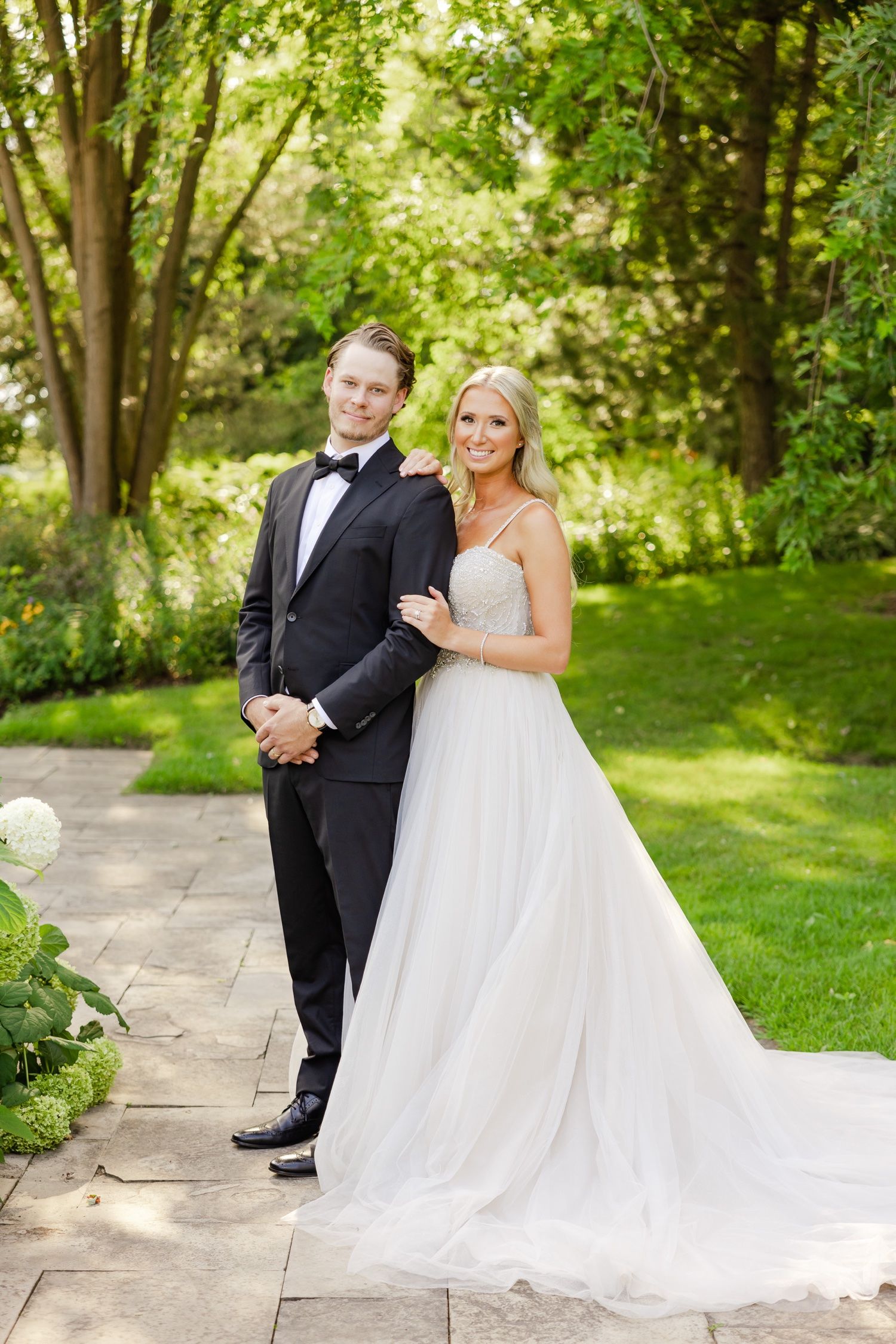 Elegant bridal couple stands on stone path with garden greenery and white flowers.