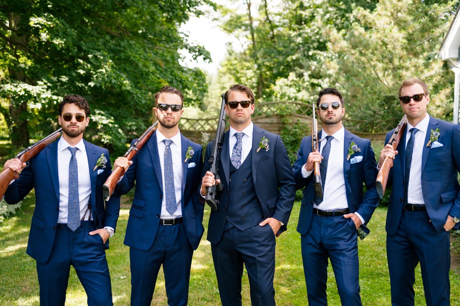 Groomsmen wearing sunglasses strike casual poses in navy suits outdoors.