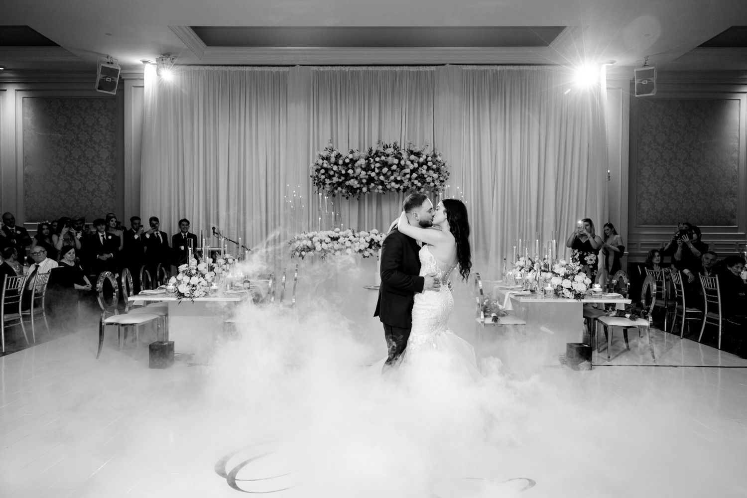 Black and white photo of a wedding dance in a beautifully decorated ballroom filled with fog effects.