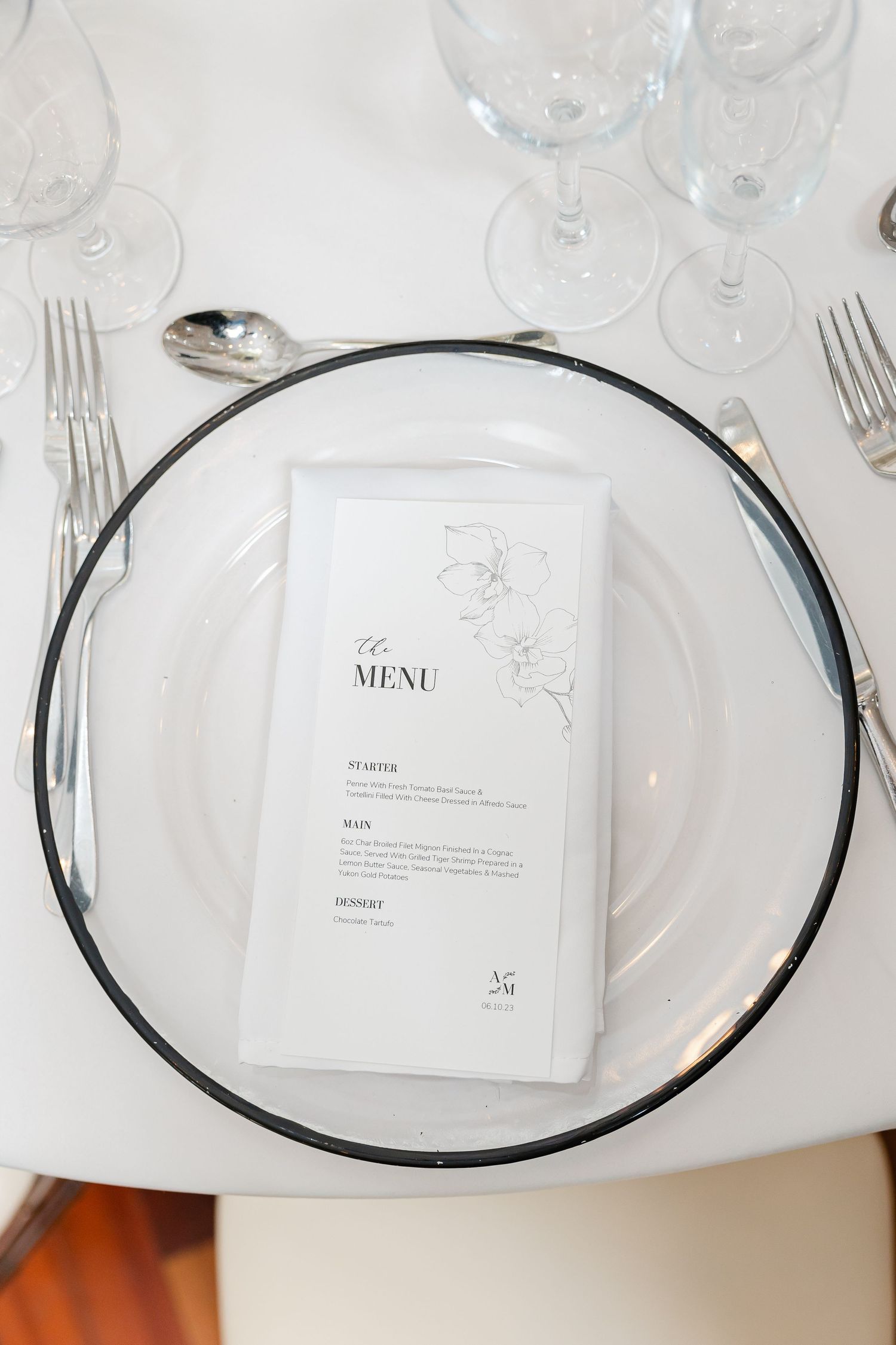 Wedding place setting with clear charger plate, menu card, and wine glasses on white tablecloth.
