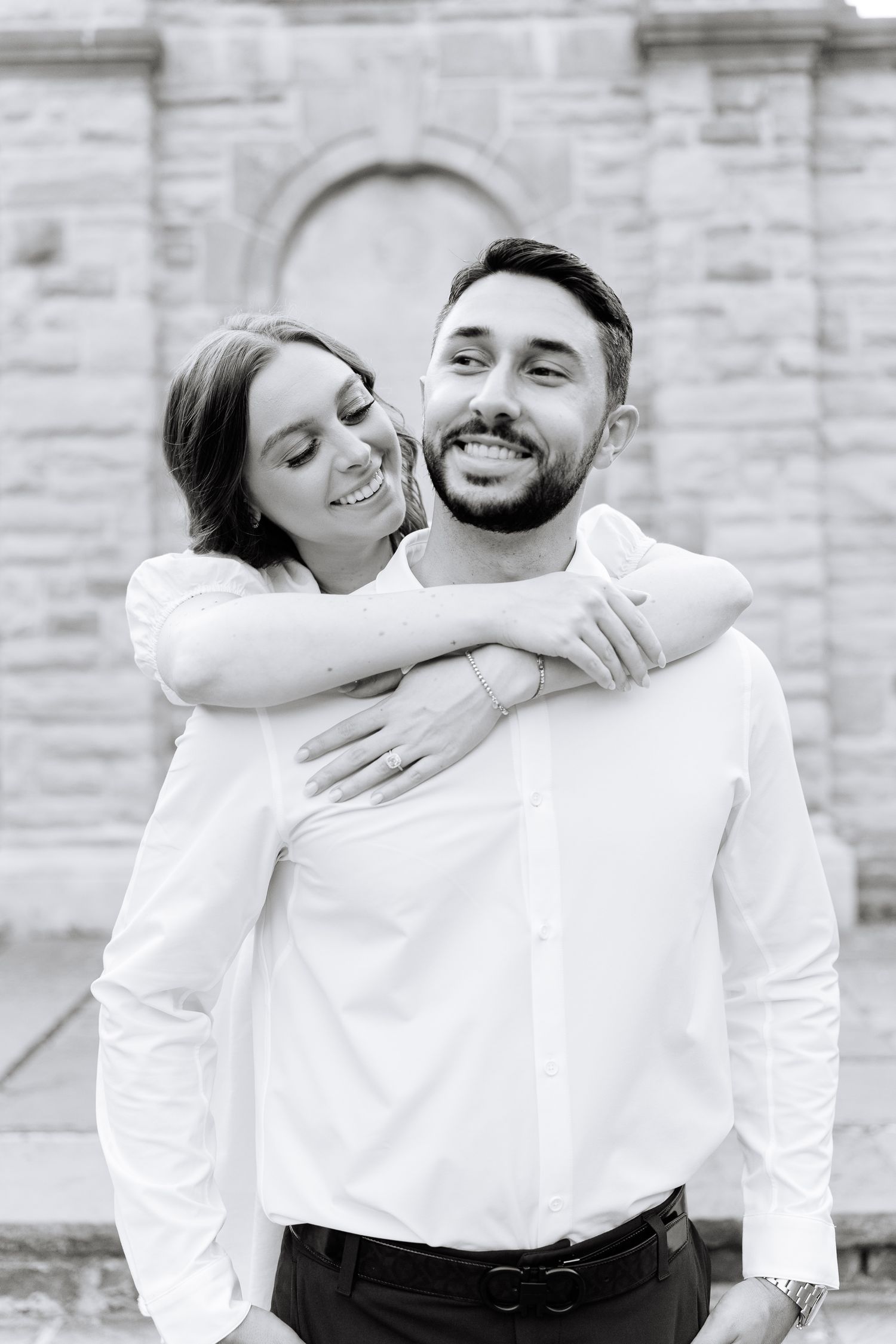 A black and white portrait of a couple embracing and smiling against a stone wall background.
