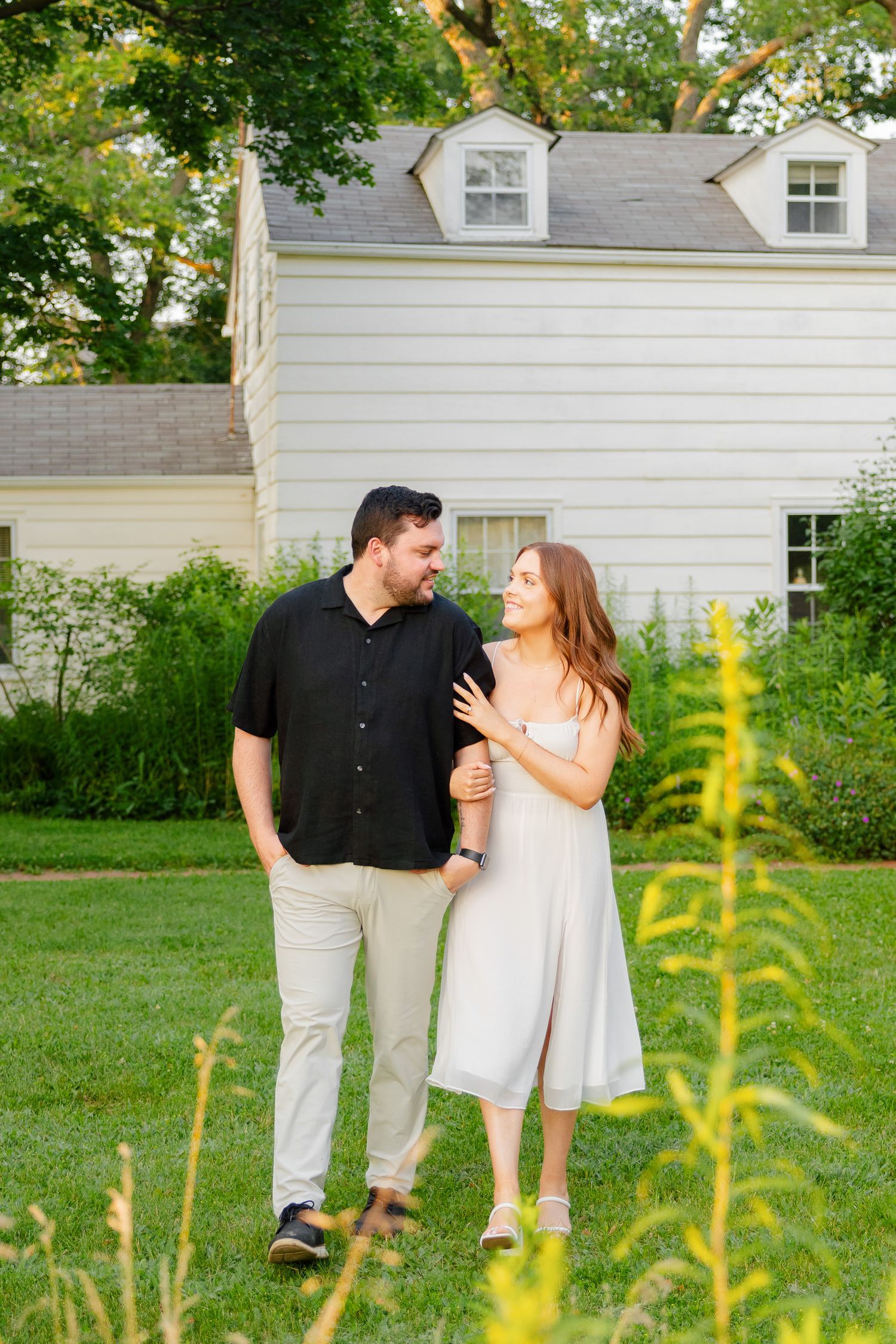 Couple sharing affectionate moment on lawn with white house and golden wildflowers in foreground.