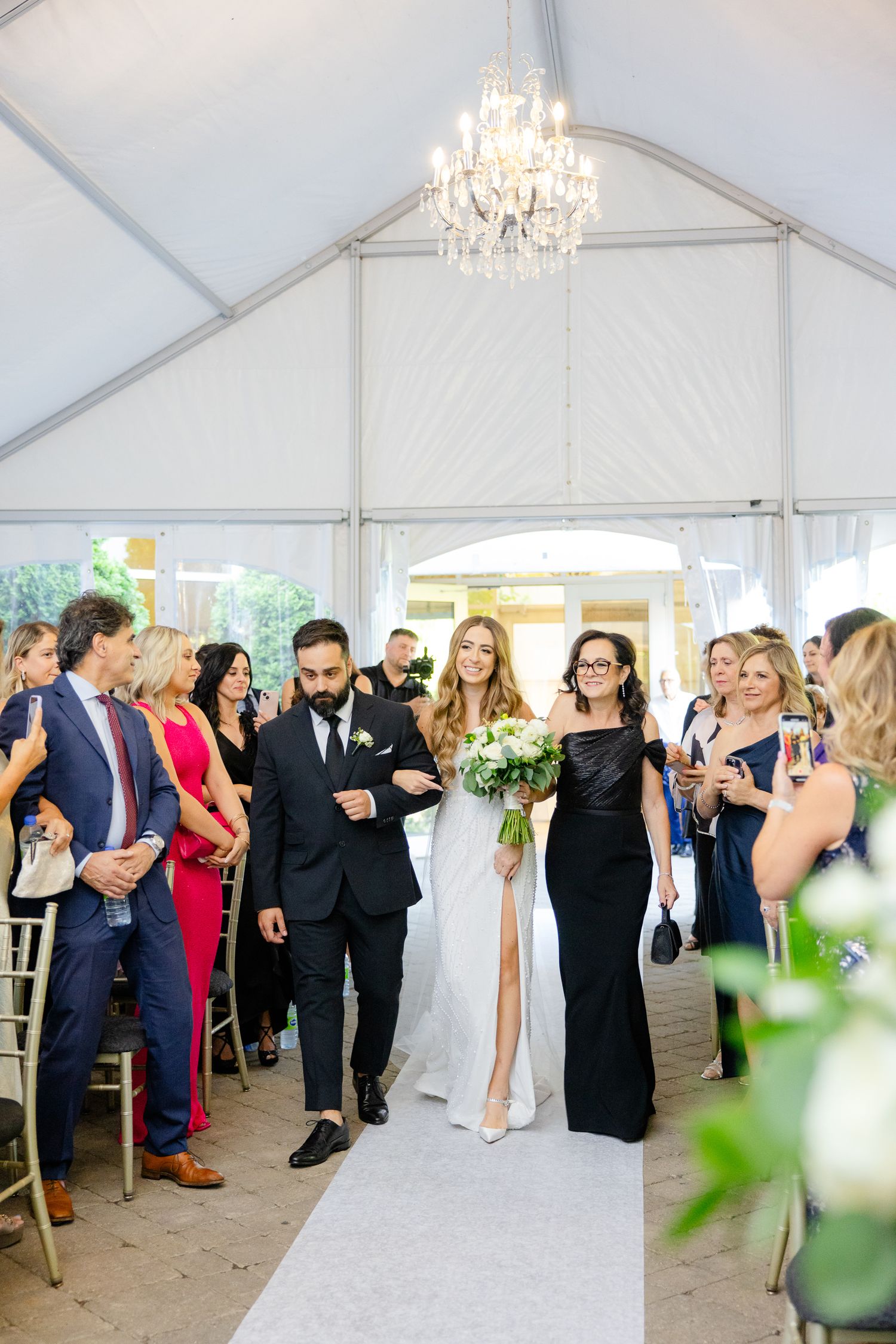 Wedding processional under crystal chandelier with guests watching from chiavari chairs.