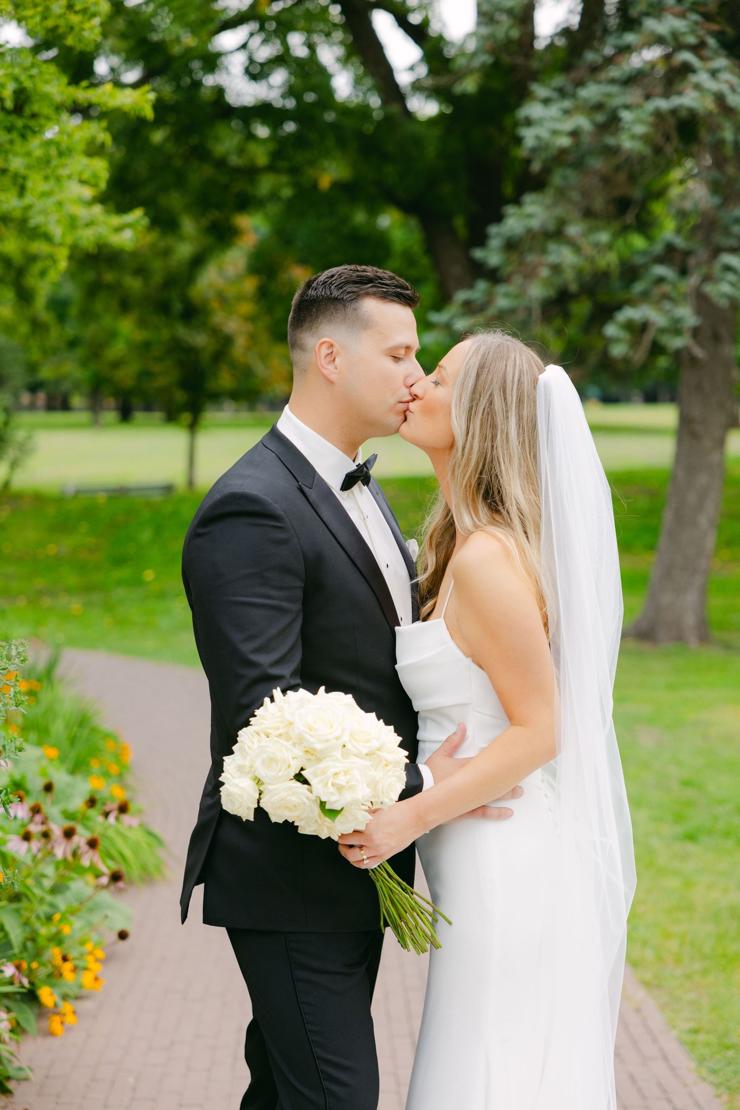 A wedding couple shares a kiss in a garden setting while holding a white bouquet.