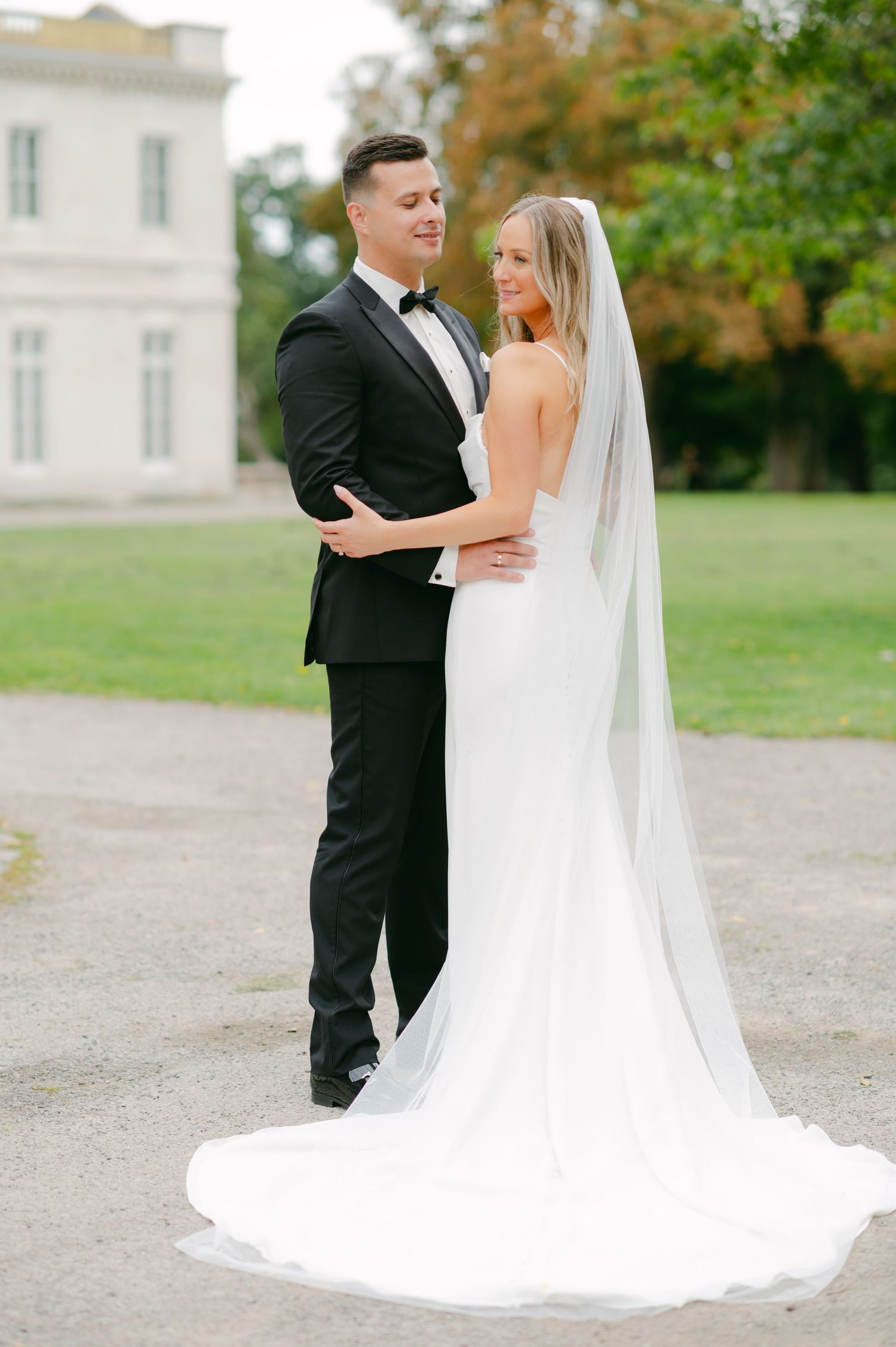 Elegant wedding portrait with white mansion backdrop and flowing bridal train.
