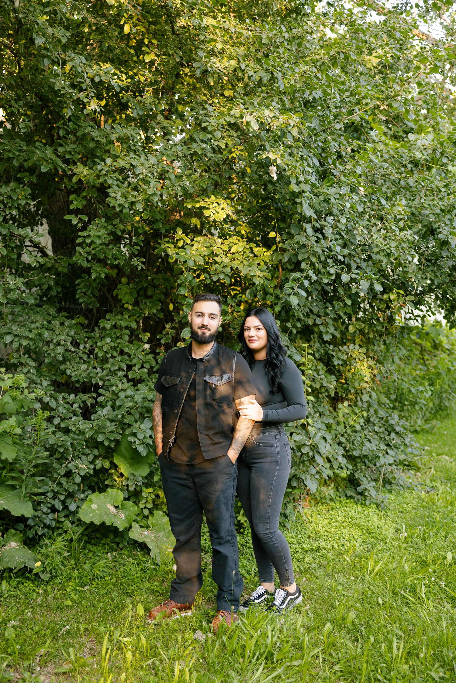 A couple in casual attire poses together against a backdrop of green foliage in a park.