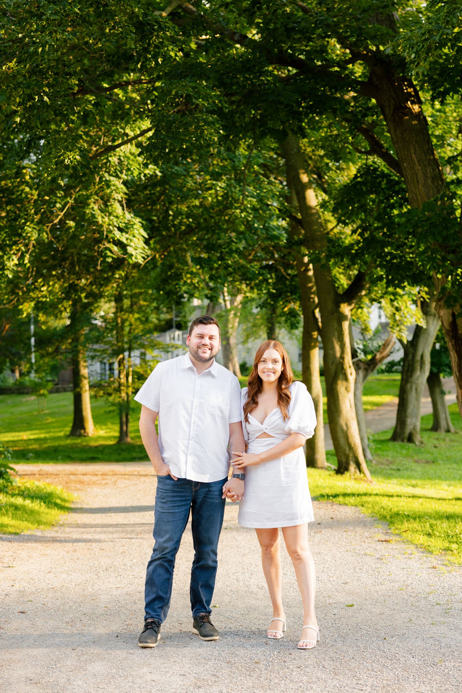 A couple poses on a tree-lined path in a sunlit park during golden hour.
