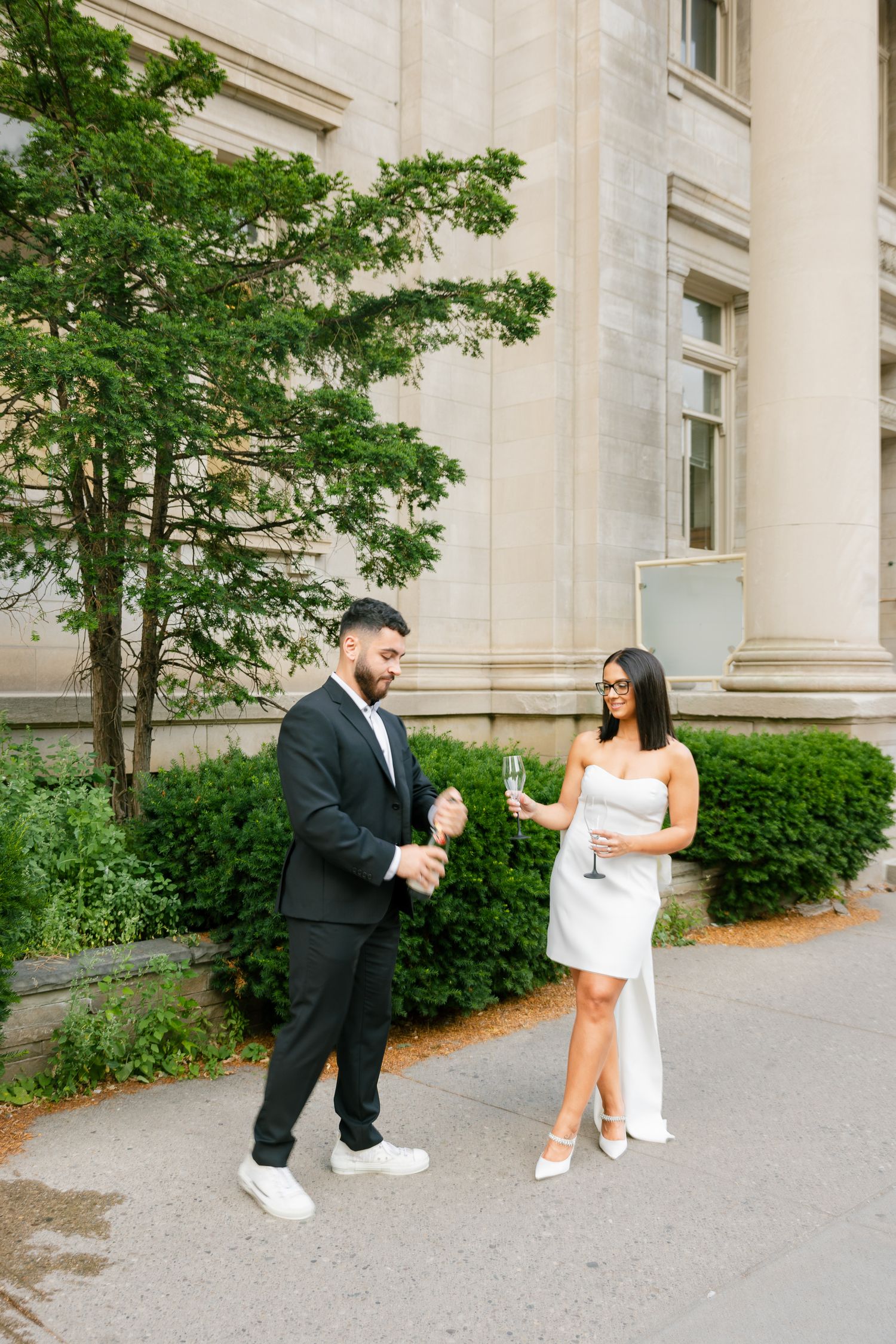 A couple in contrasting black and white outfits share a playful moment outside a classic stone building.
