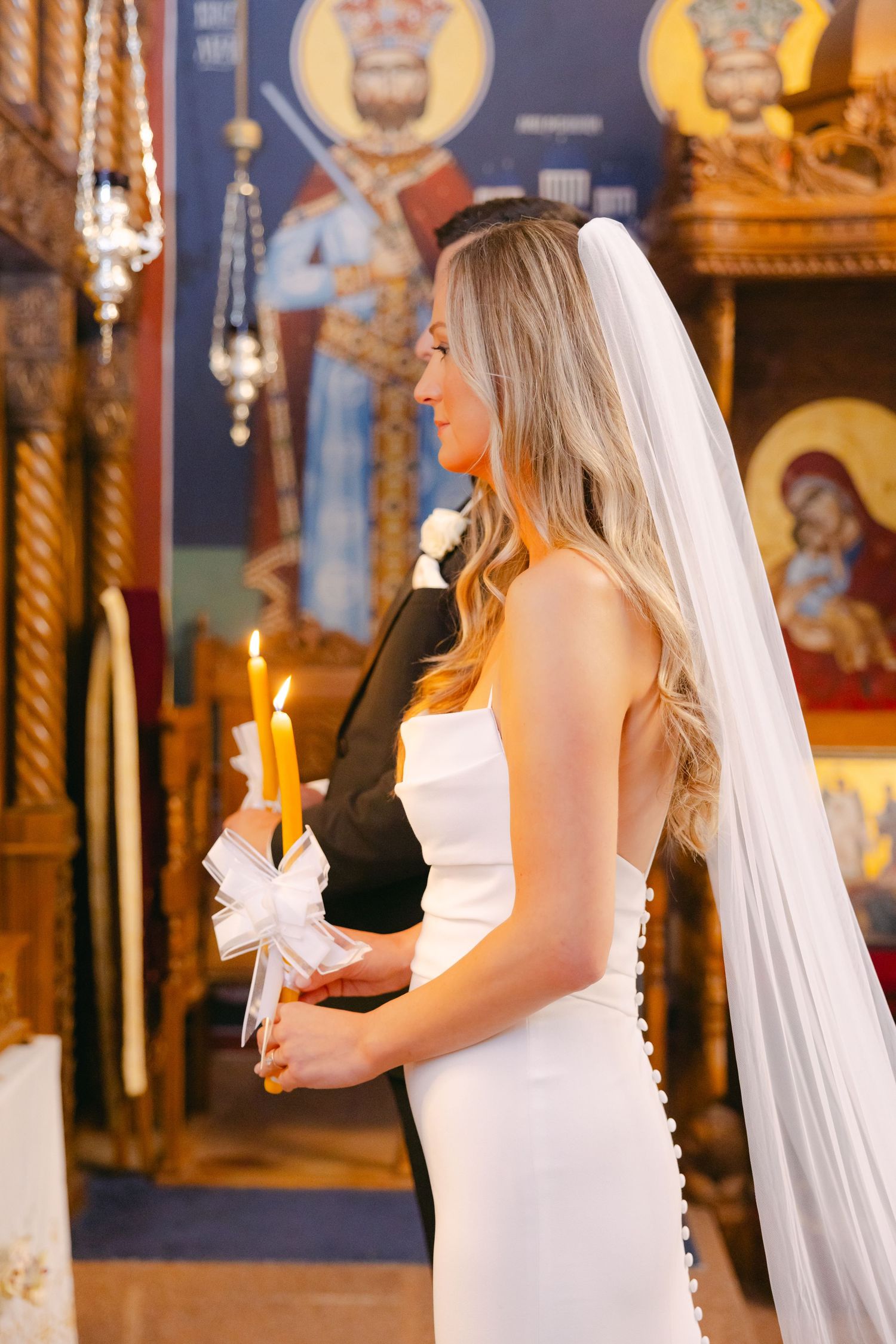 Bride holding ceremonial candle during Orthodox wedding ceremony surrounded by religious artwork.