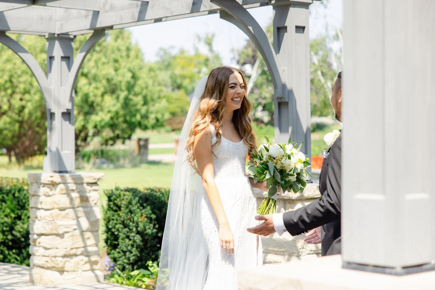 Elegant wedding moment captured under a white pergola in a manicured garden setting.