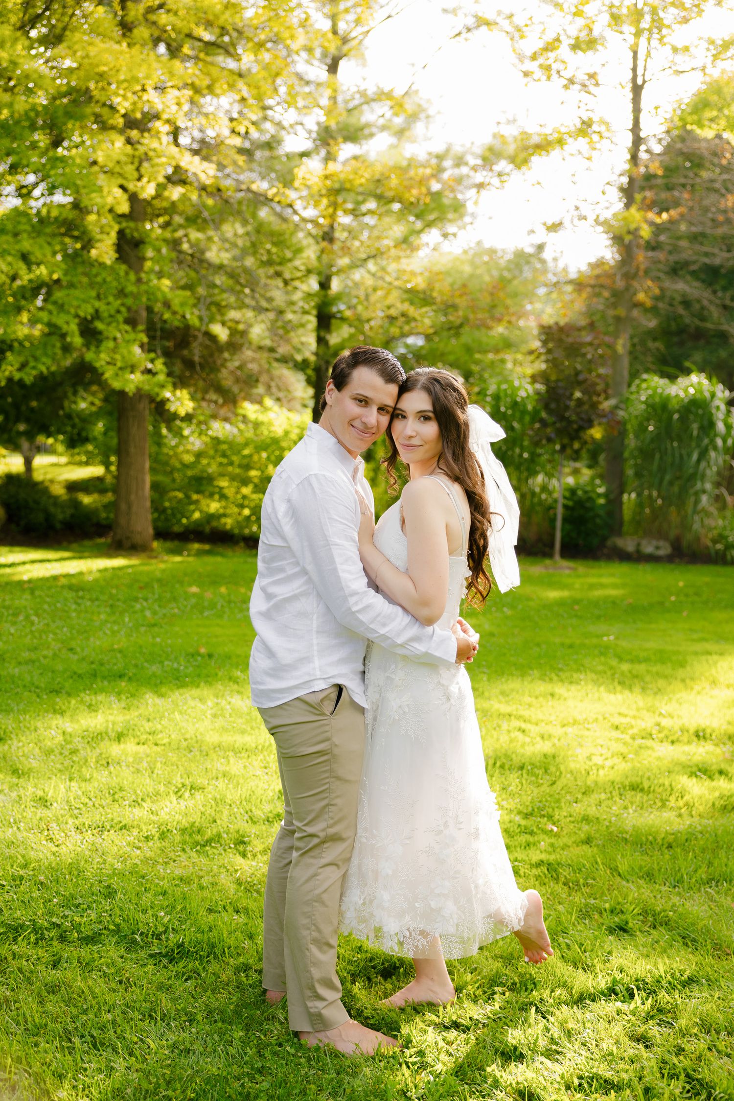 Casual wedding portrait of a couple standing barefoot on grass in a sunlit garden.