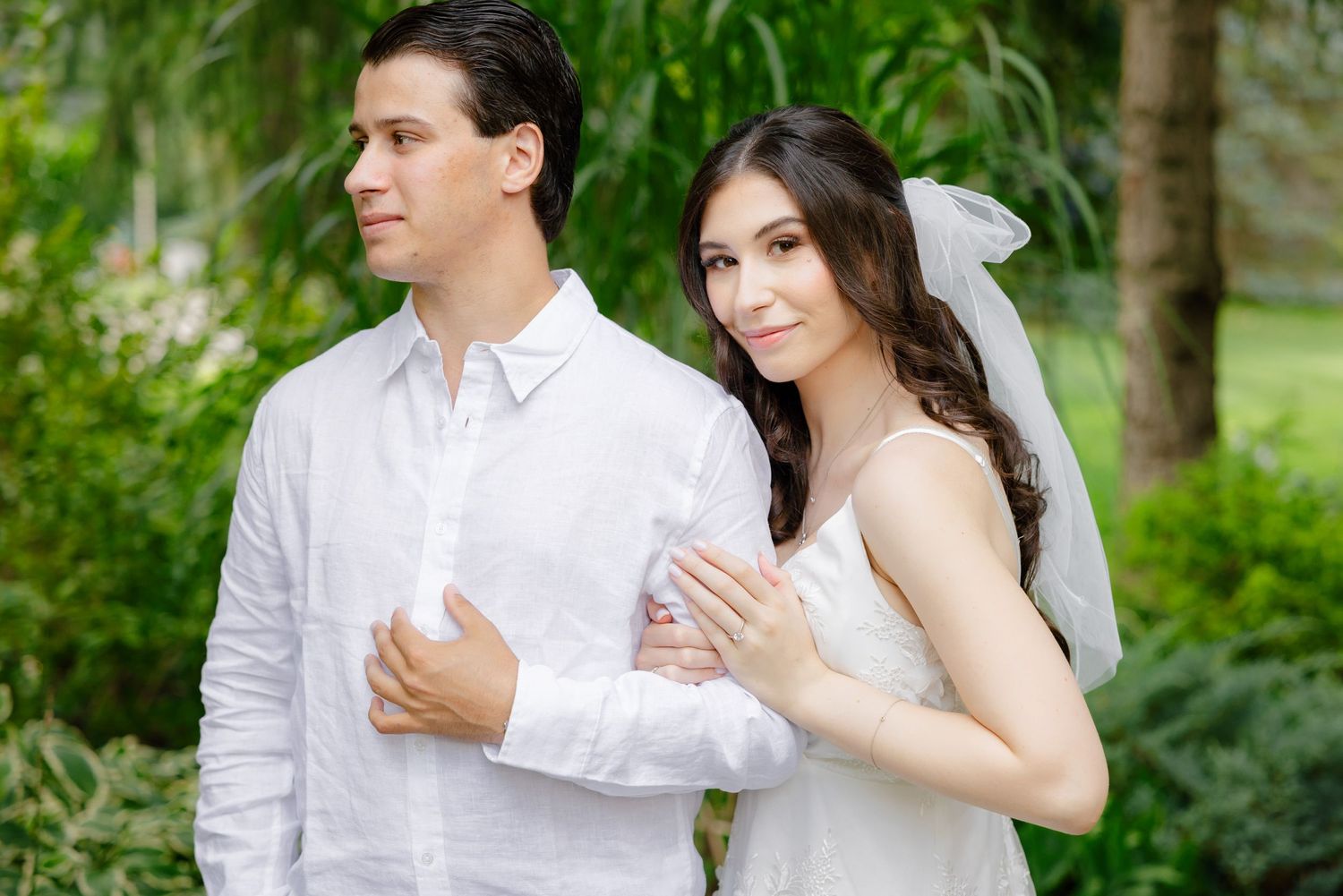 A couple in wedding attire embraces outdoors in a garden setting under evergreen trees.