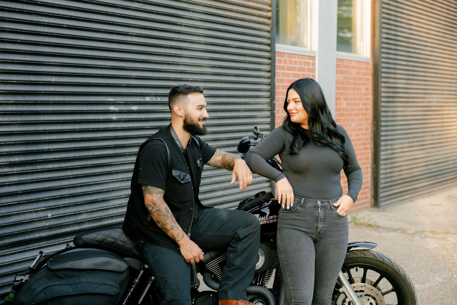 Urban couple posing casually next to motorcycle against industrial backdrop.