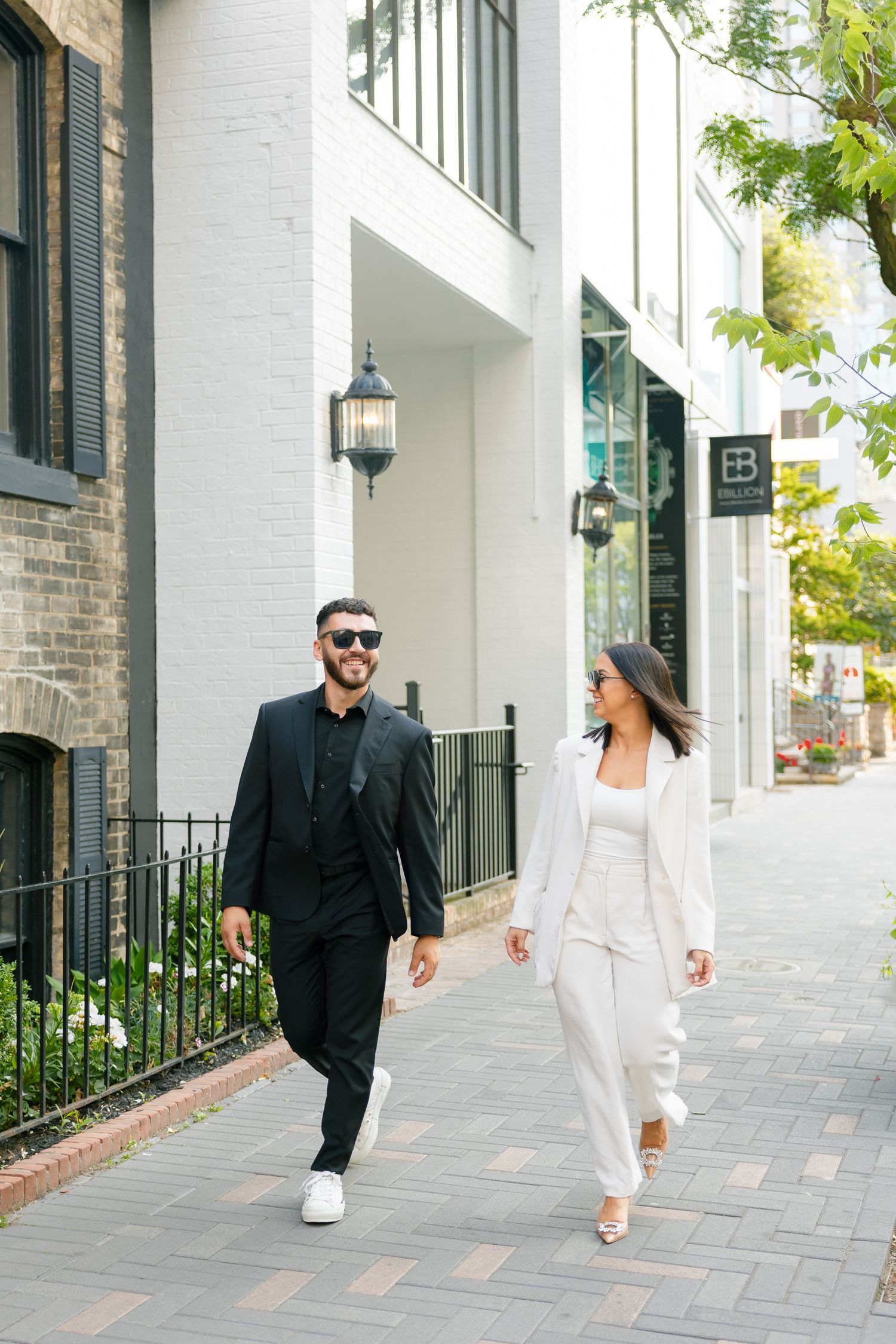 A stylishly dressed couple walks along an urban sidewalk with white buildings and lanterns.