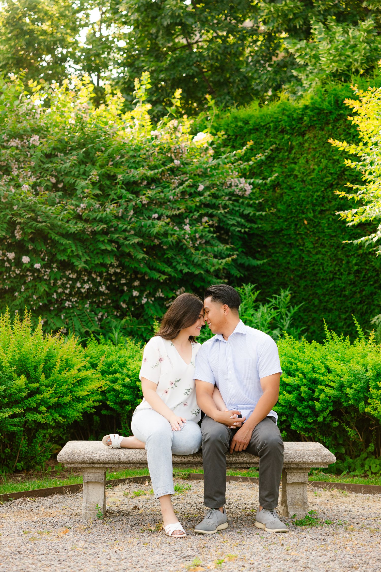 A couple sits close together on a stone bench surrounded by manicured hedges and flowering plants.