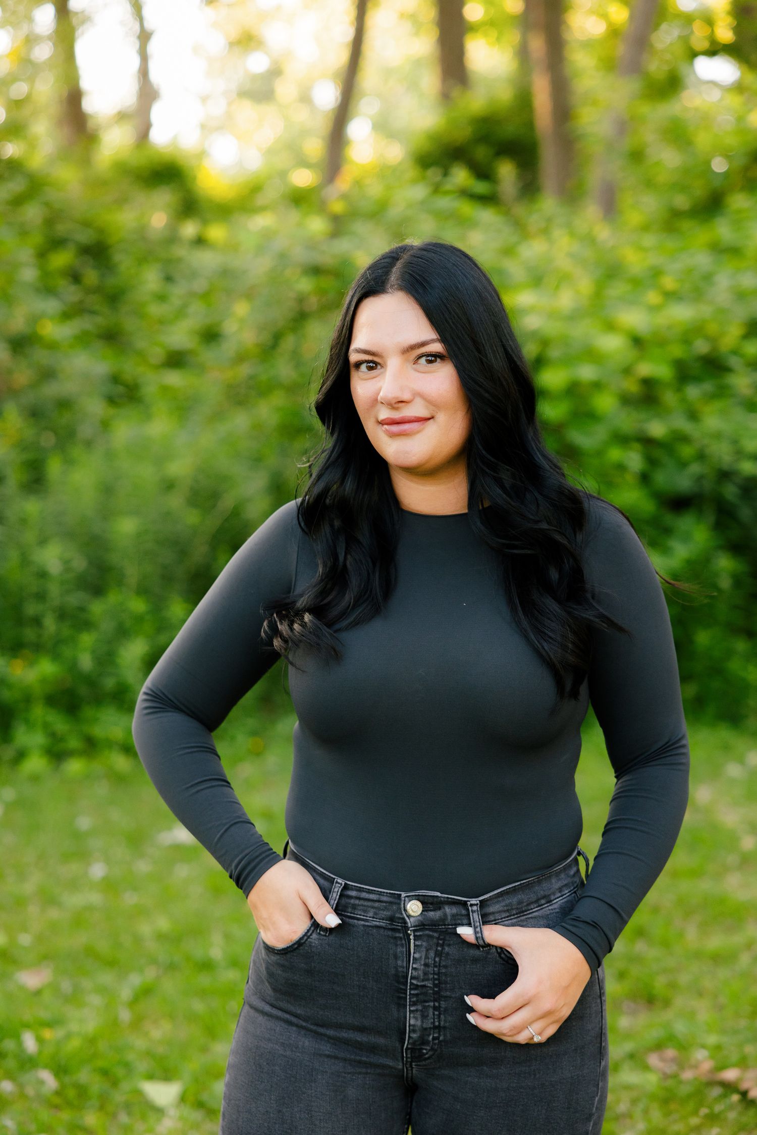 Woman in dark fitted top smiles warmly while posing in outdoor forest setting.