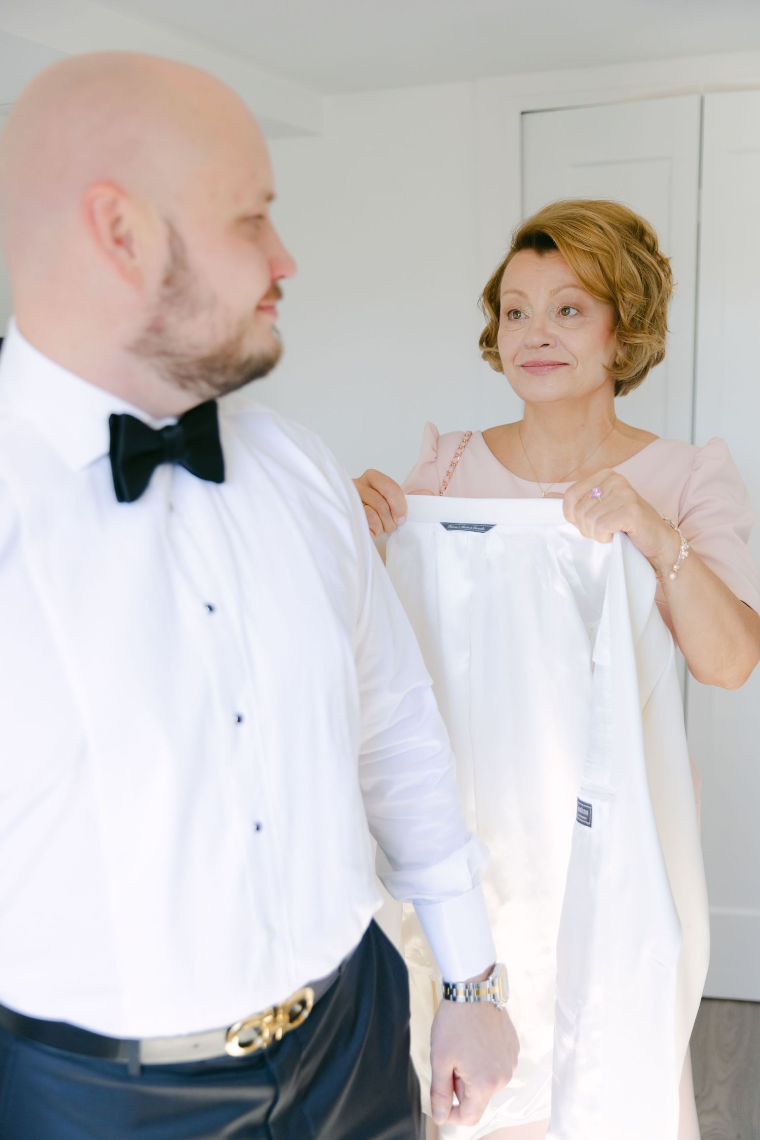 Two people in formal attire preparing for an event in a bright room.