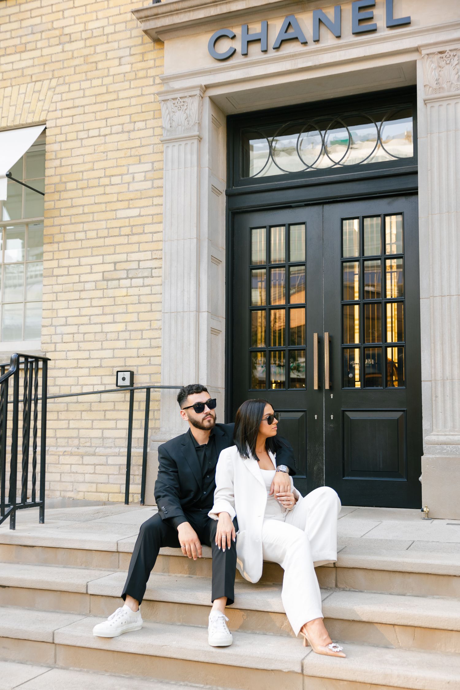 Couple in contrasting black and white outfits sitting on steps of Chanel boutique entrance.