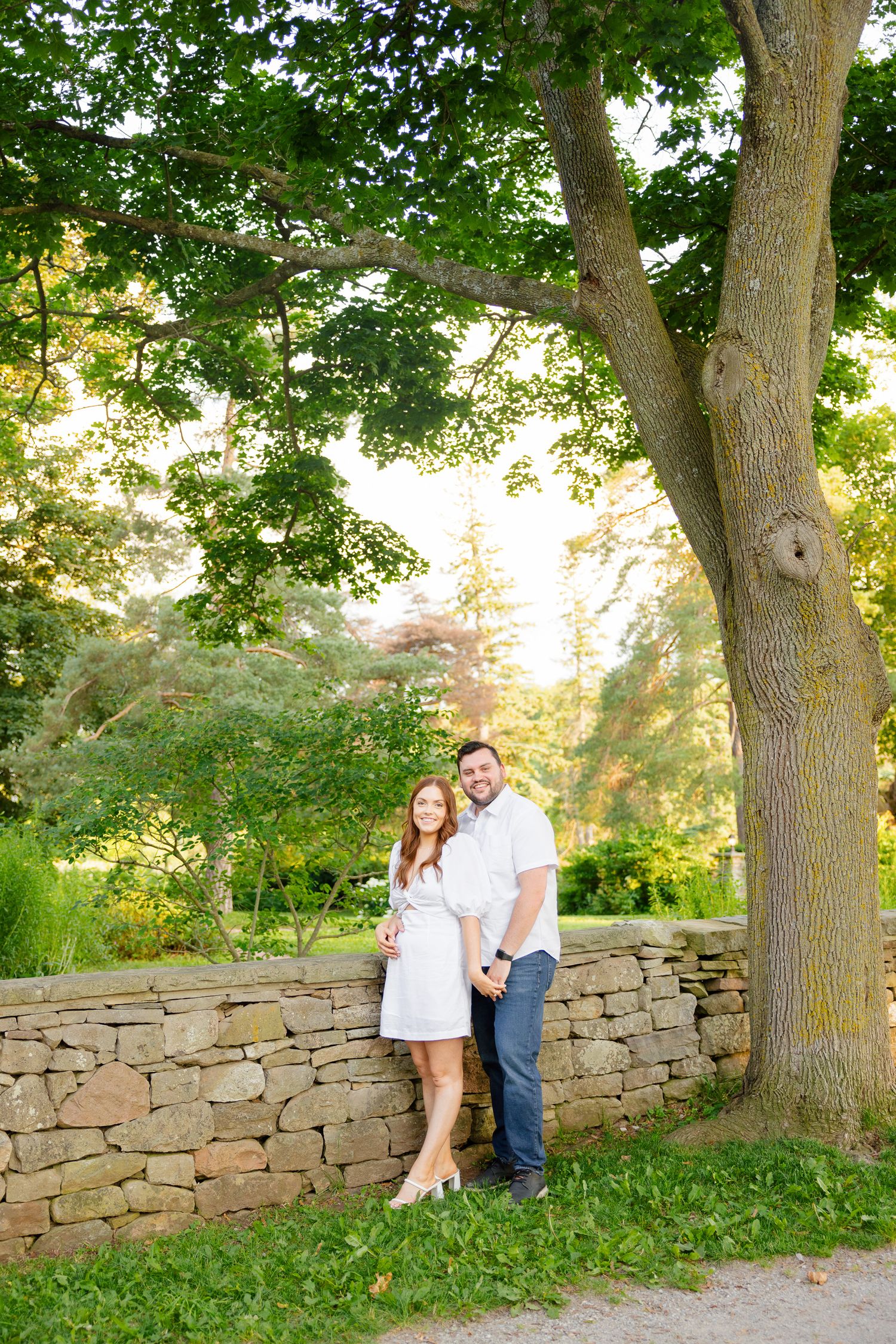 Couple in coordinating white outfits poses by stone wall in garden with glowing sunset light.