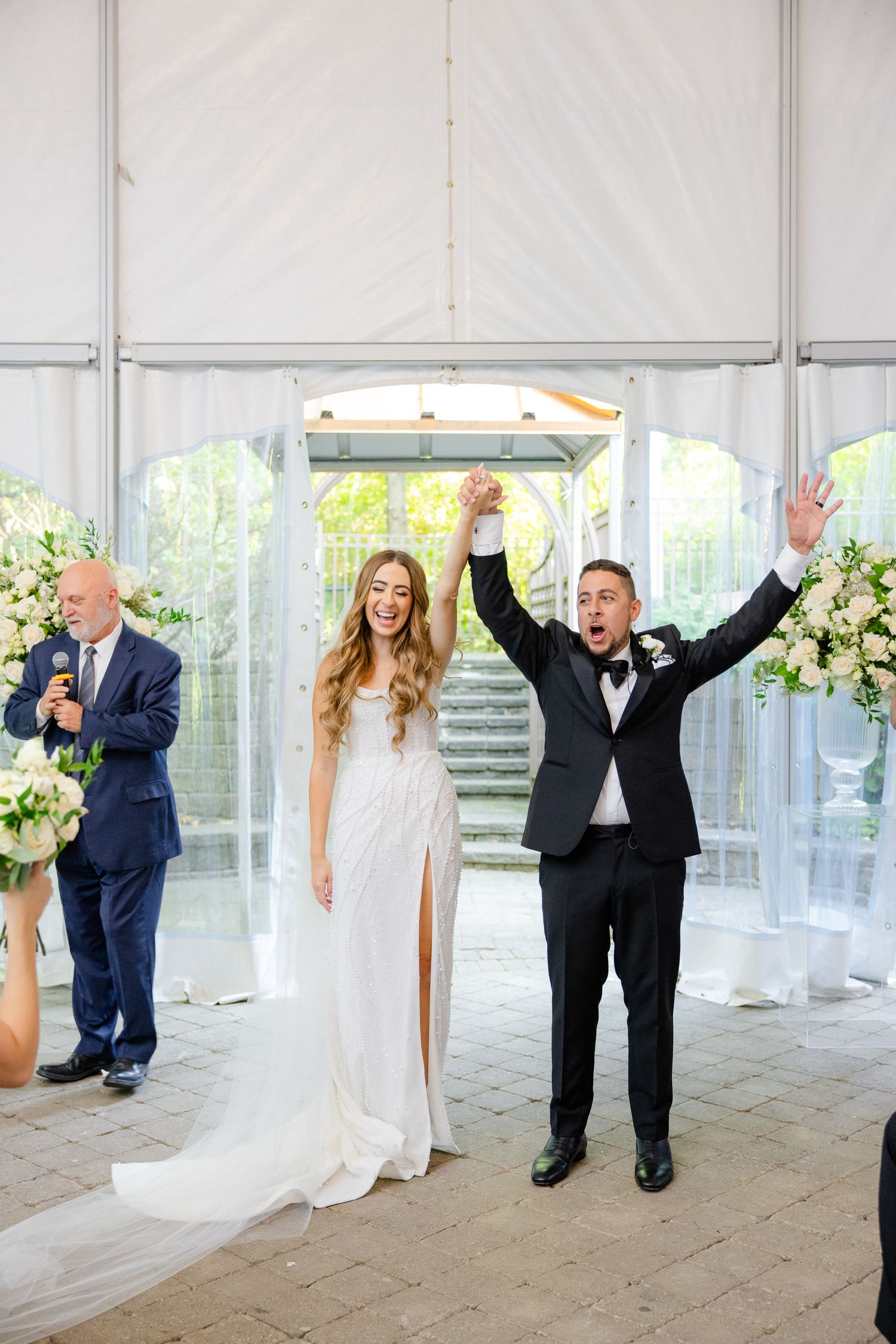 A joyful wedding ceremony captures a newlywed couple celebrating with raised arms in a white tent venue with floral decorations.