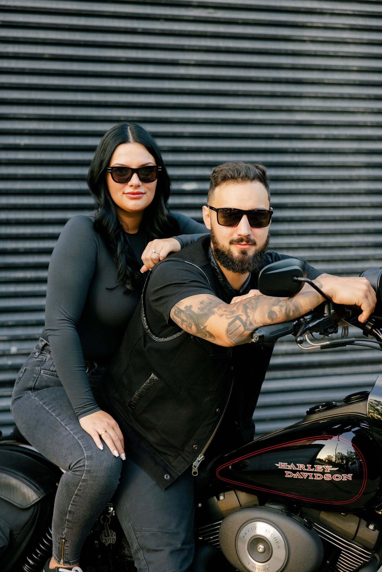 Modern couple in dark clothing poses with Harley Davidson motorcycle outdoors.