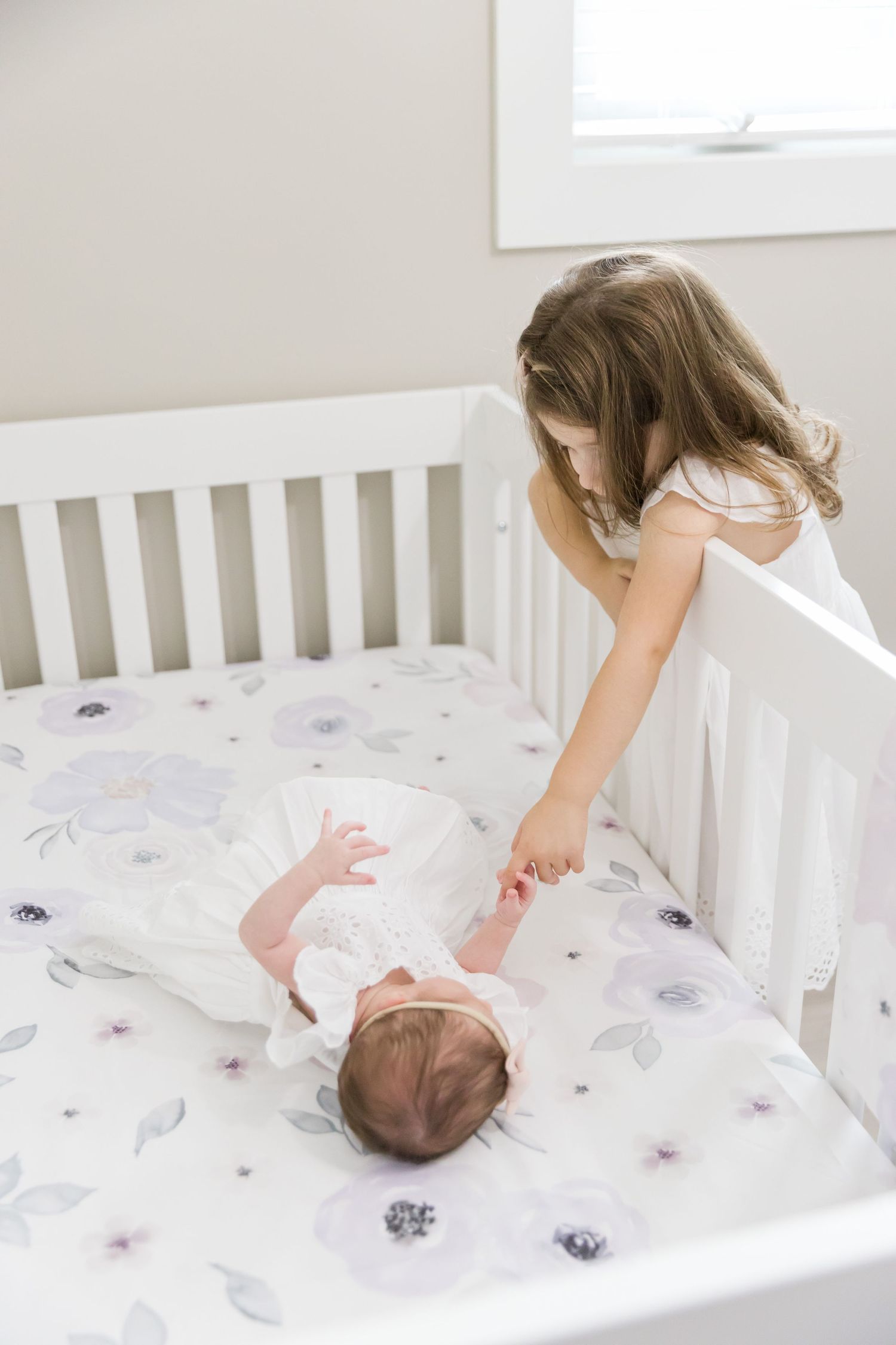 Child reaches over white crib railing to touch newborn on floral bedding.