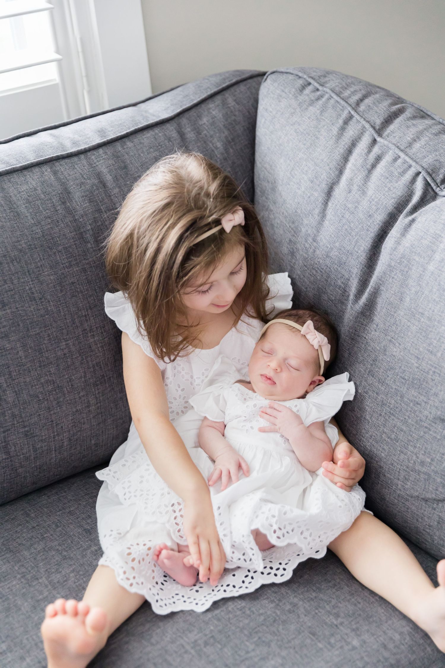 Child and sleeping baby in matching white dresses rest together on grey sofa.