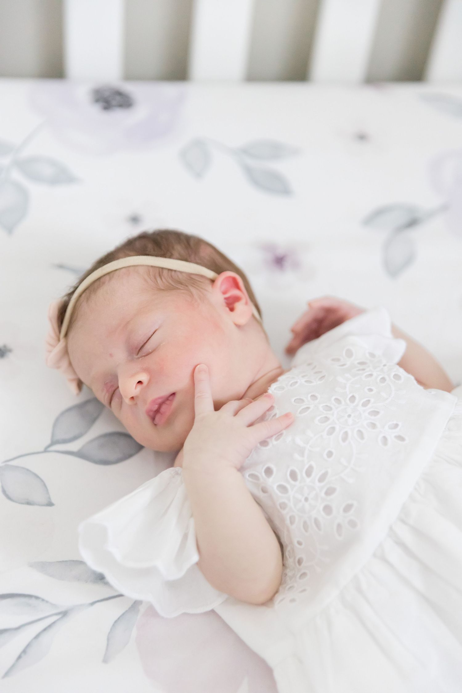 Sleeping baby wearing white eyelet lace dress rests hand on cheek while lying on floral bedding.