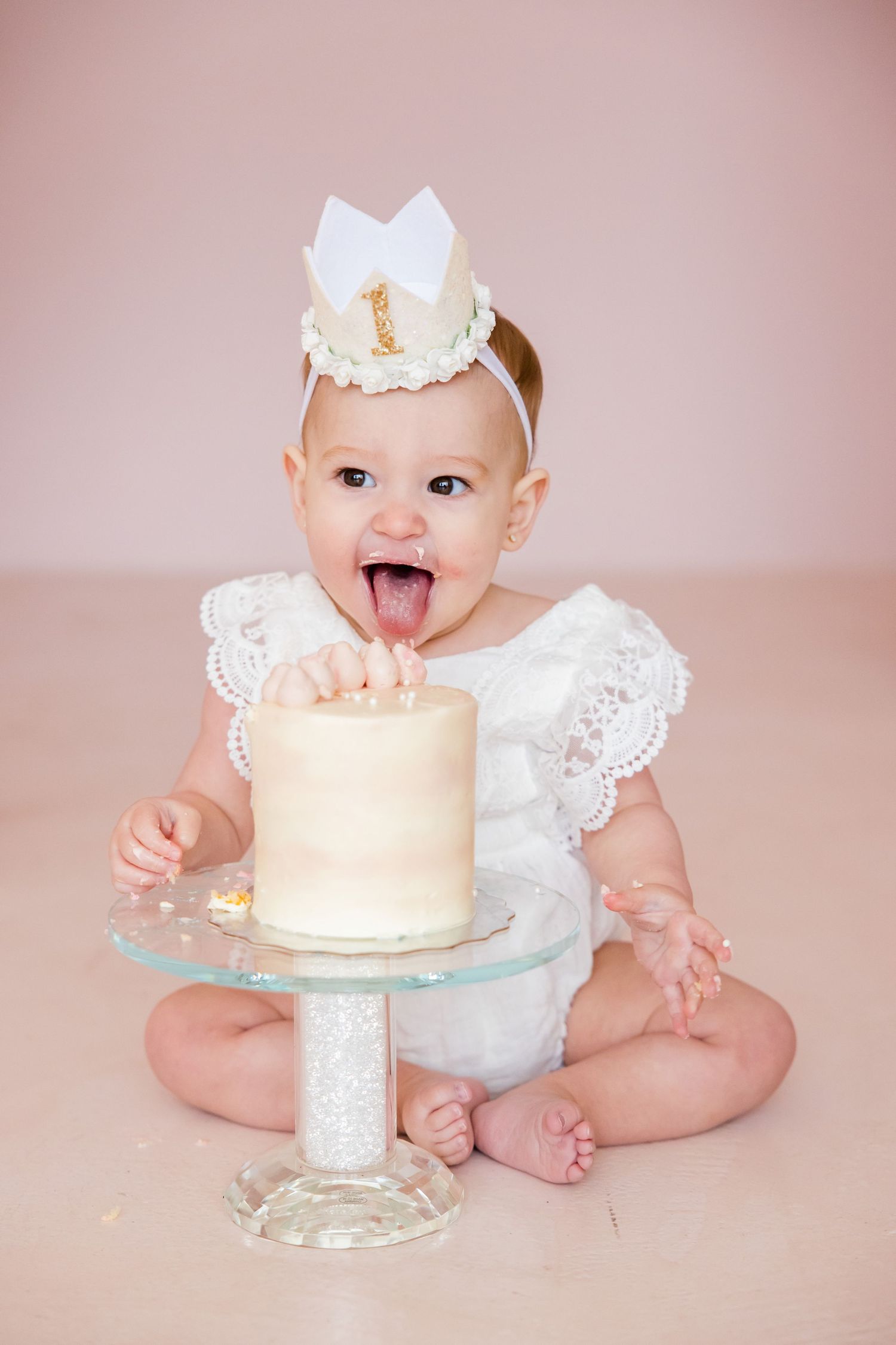 First birthday cake smash portrait with white dress and crown against pink backdrop.