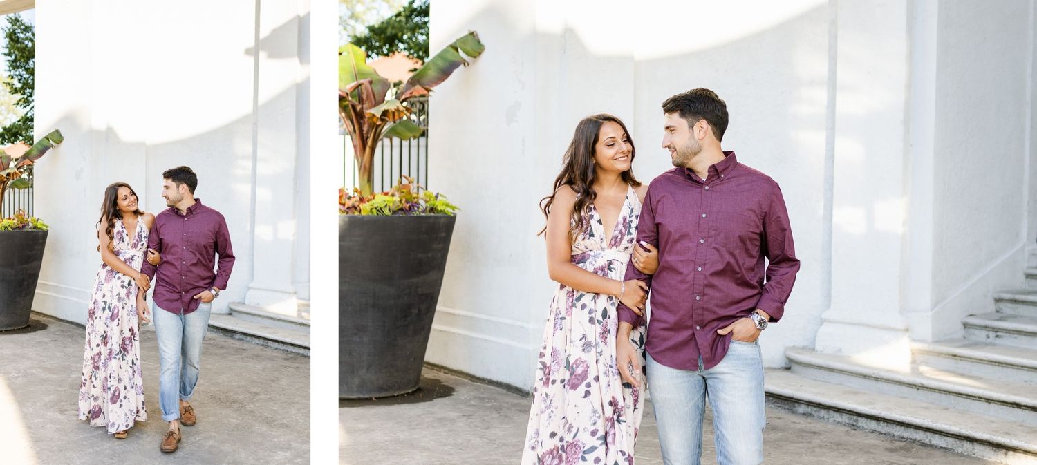 A couple walks together along a white columned walkway, with her in a floral dress and him in burgundy.
