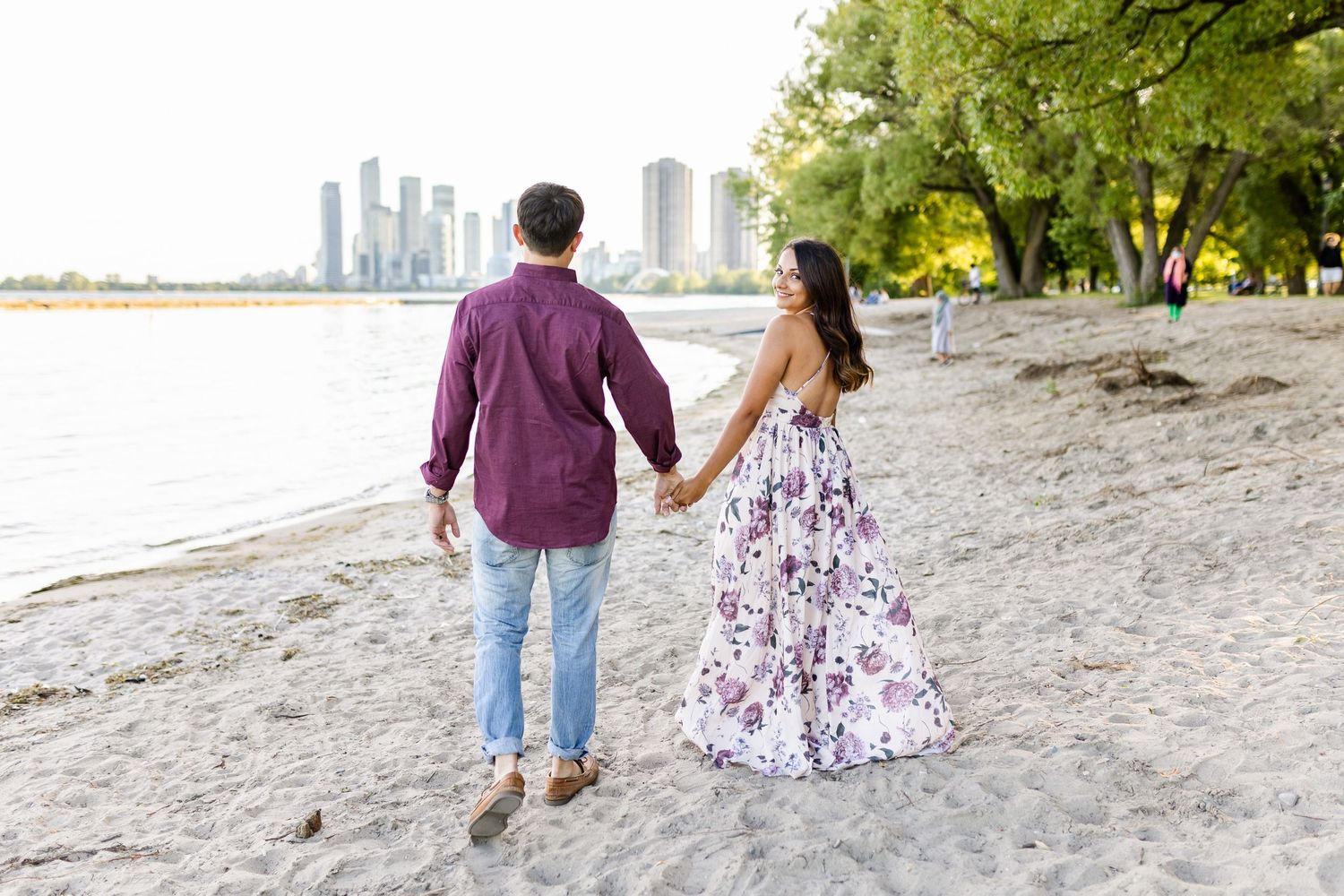 Couple strolling along sandy beach with urban skyline visible in distance.