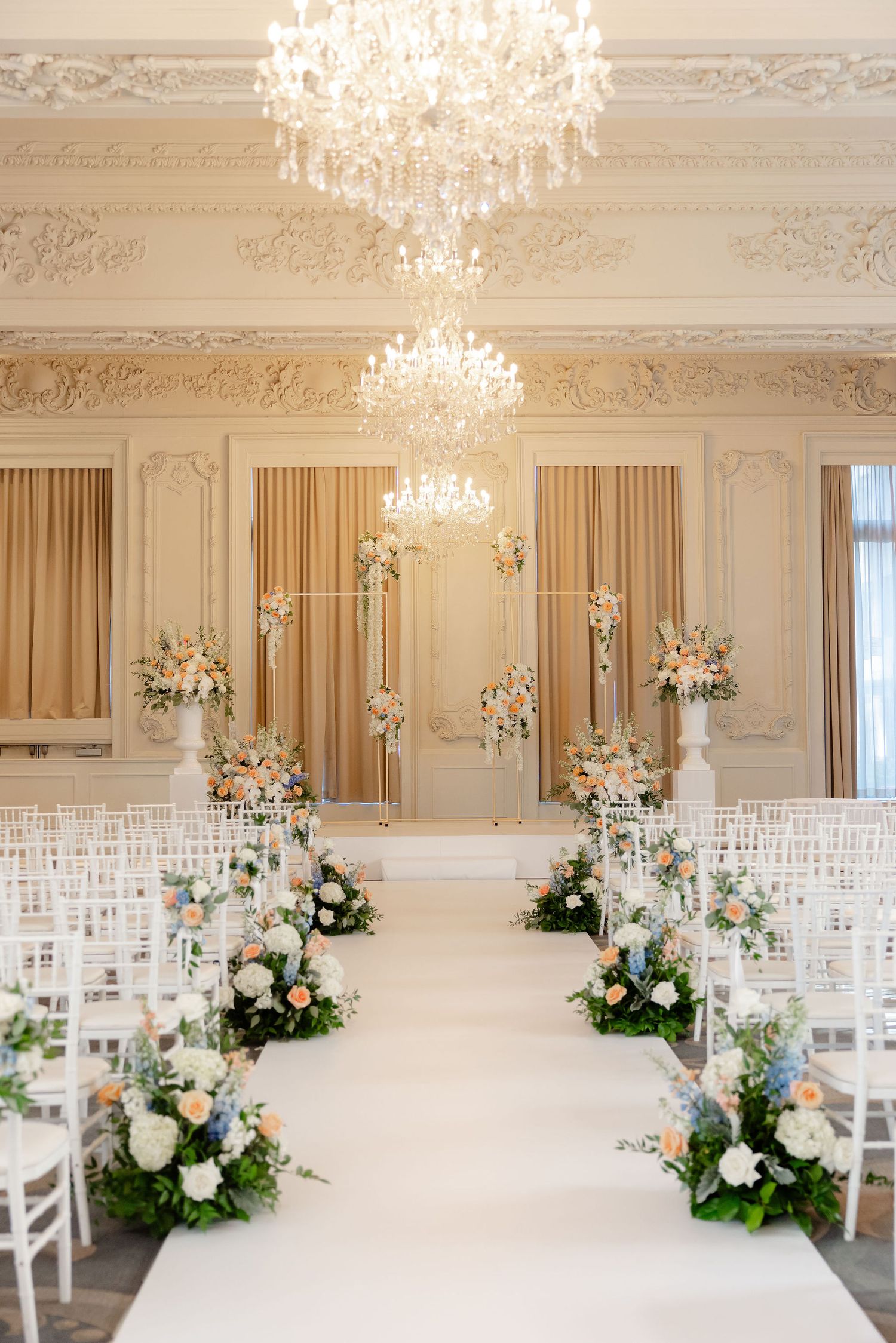 Elegant wedding ceremony setup with white chairs, crystal chandeliers, and floral arrangements in a luxurious cream-colored room.