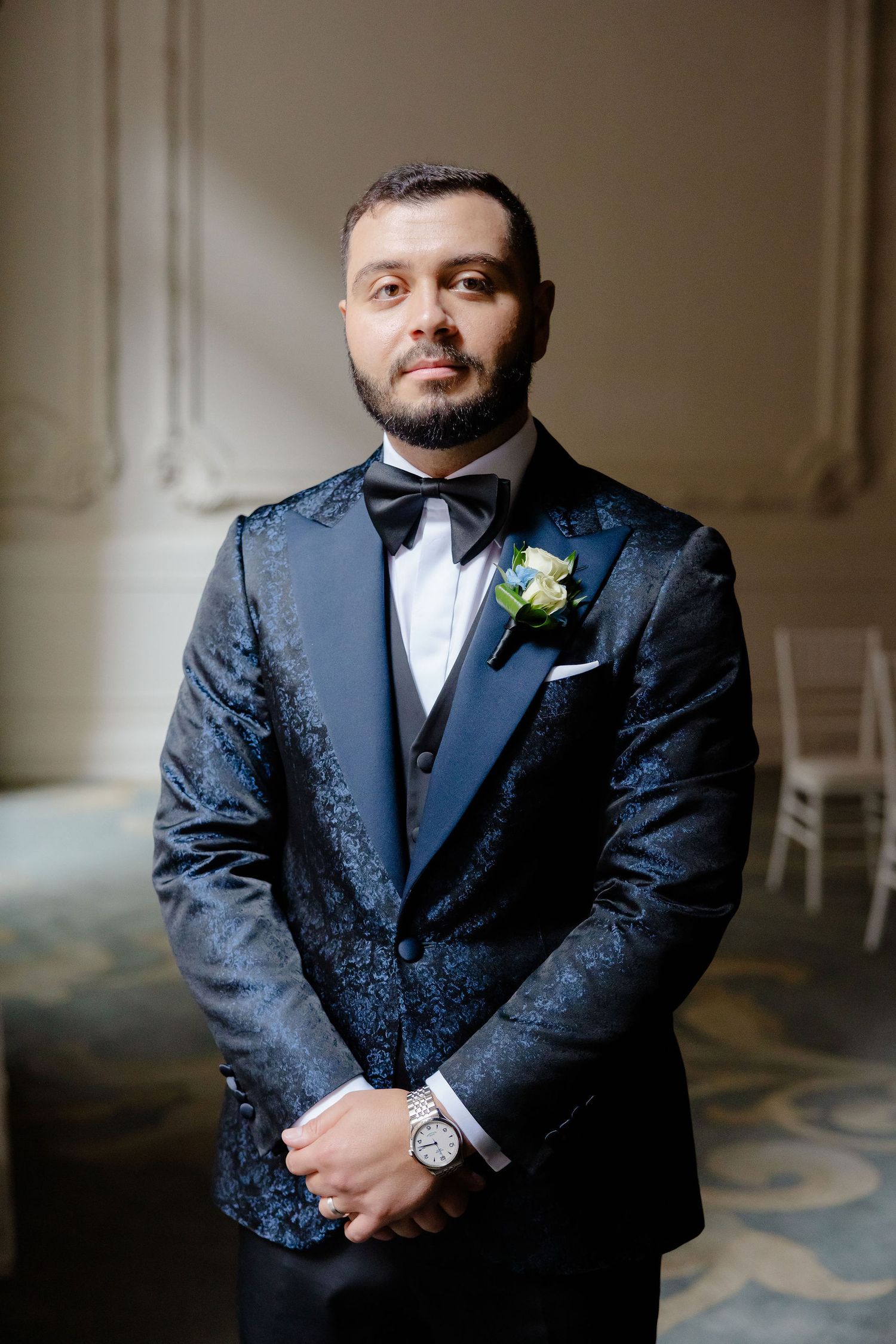 A groom in a navy blue patterned tuxedo with bowtie and boutonniere poses for a formal wedding portrait.