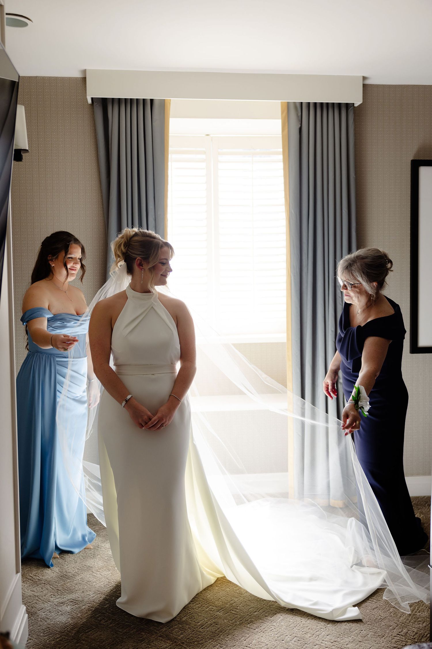 Wedding preparation scene with a bride in a sleek white gown being attended to by two people in a hotel room.