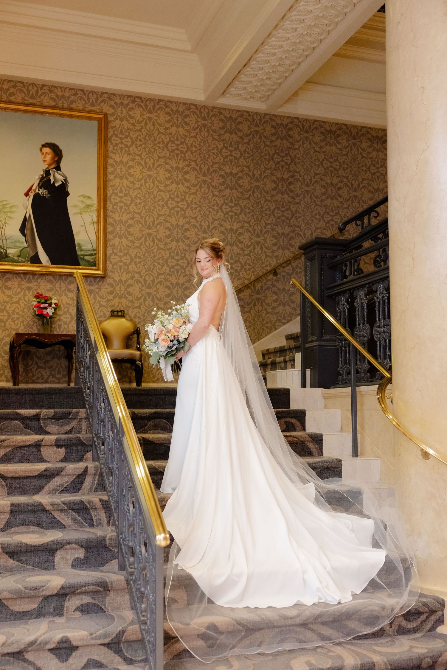Bride in flowing white gown poses on grand staircase with ornate railings and patterned carpet.