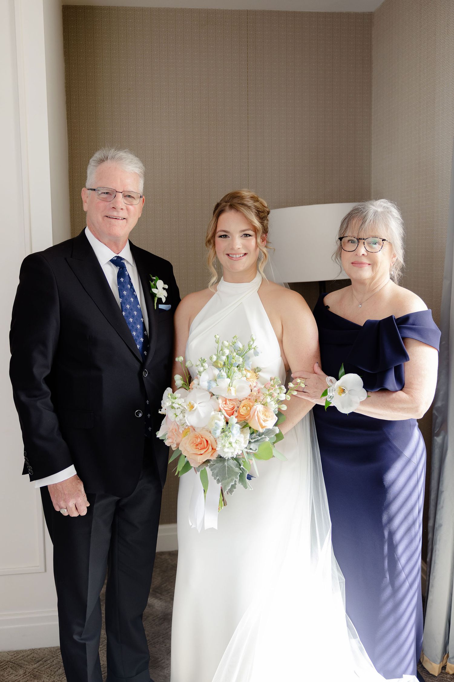 Wedding portrait featuring a bride in halter dress with peach bouquet alongside two formal-dressed companions.