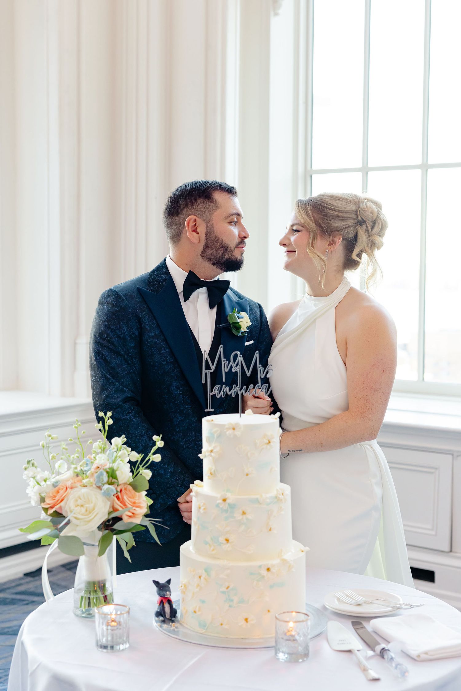 A couple shares an intimate moment beside their elegant three-tiered white wedding cake decorated with pastel accents.