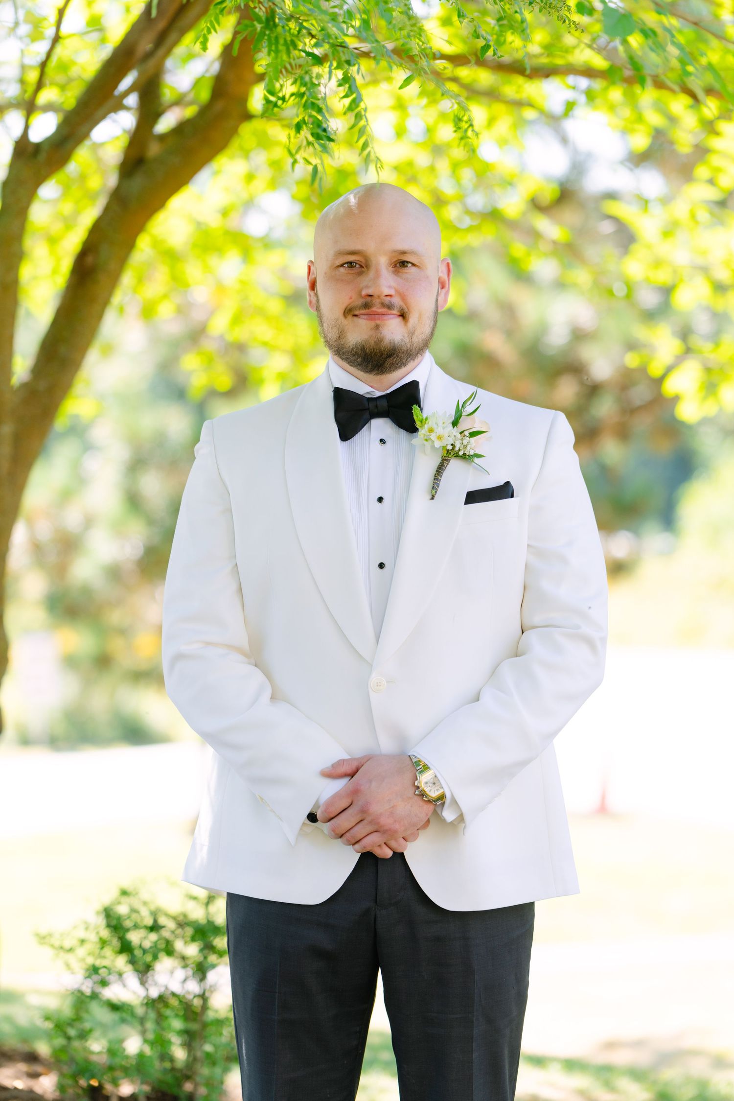 Formal portrait of groom in white tuxedo jacket and black bow tie under tree.