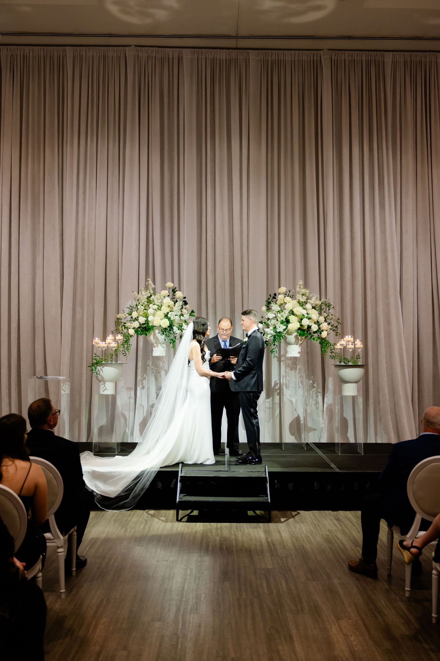 Wedding ceremony from center aisle showing dramatic long veil and elegant floral displays.