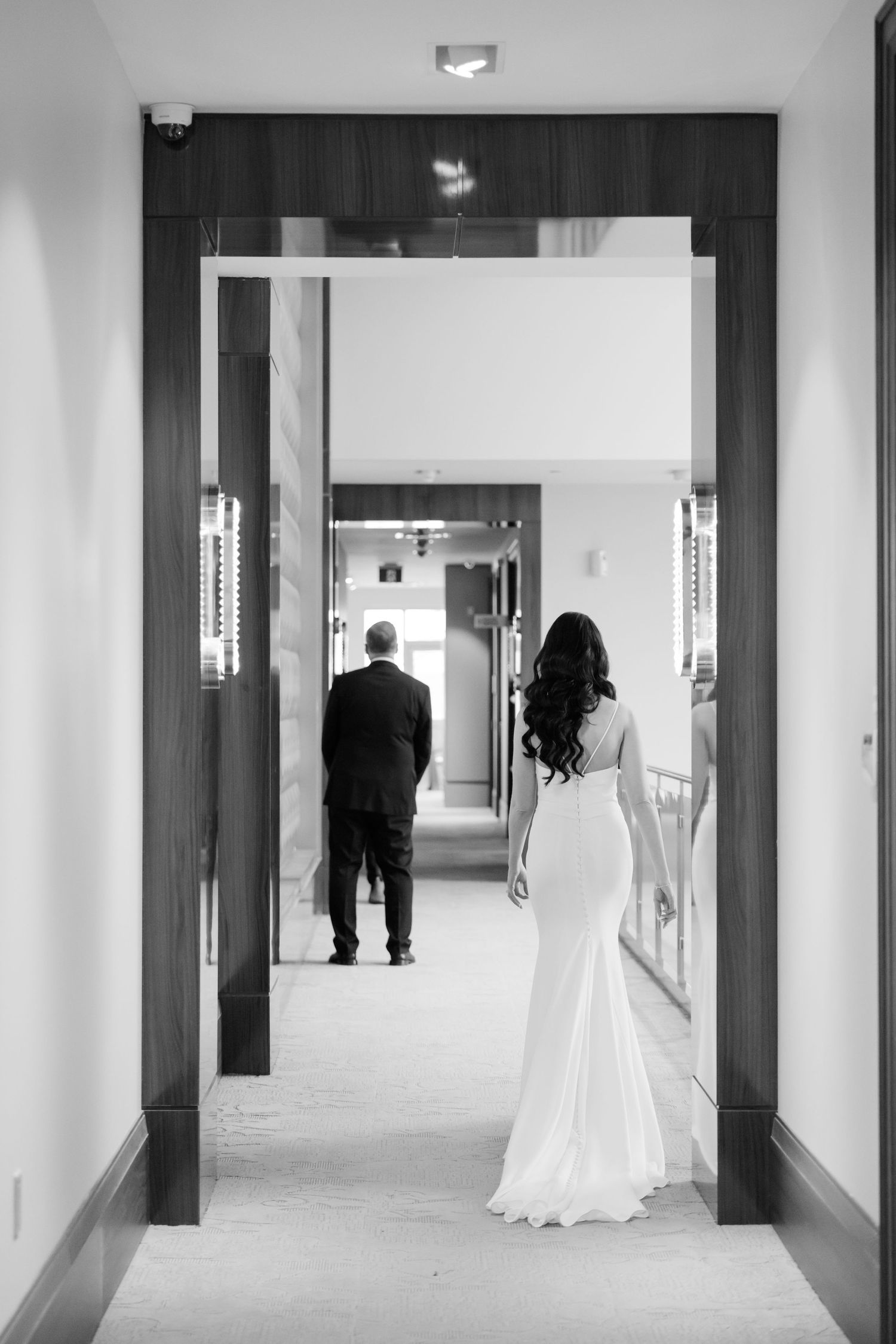 Silhouetted figures in formal attire stand at opposite ends of an elegant hallway.