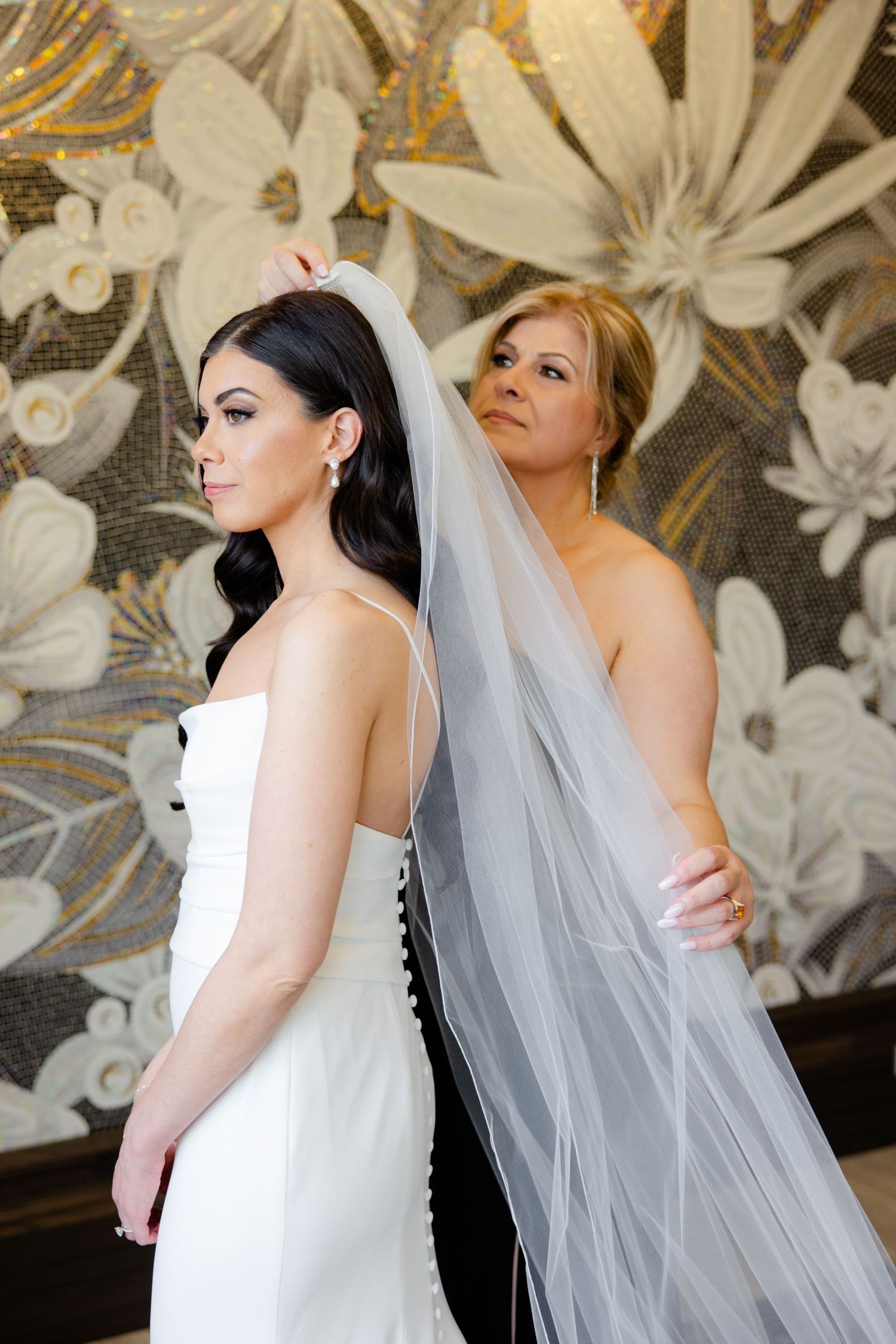 A wedding scene with flowing veil adjustments against a golden floral wallpaper.